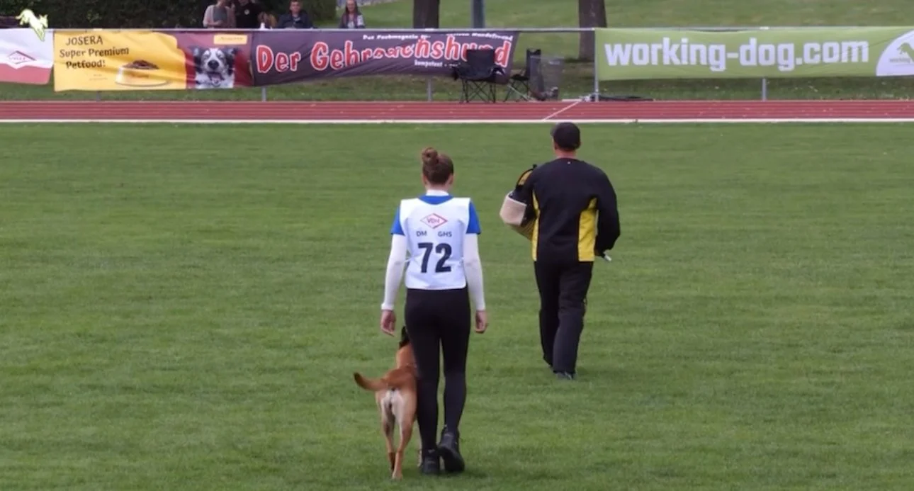 Ein Mann und eine Frau gehen über eine Sportwiese, begleitet von einem Hund. Im Hintergrund sind Banner an einer Bande und einige Zuschauer zu sehen.