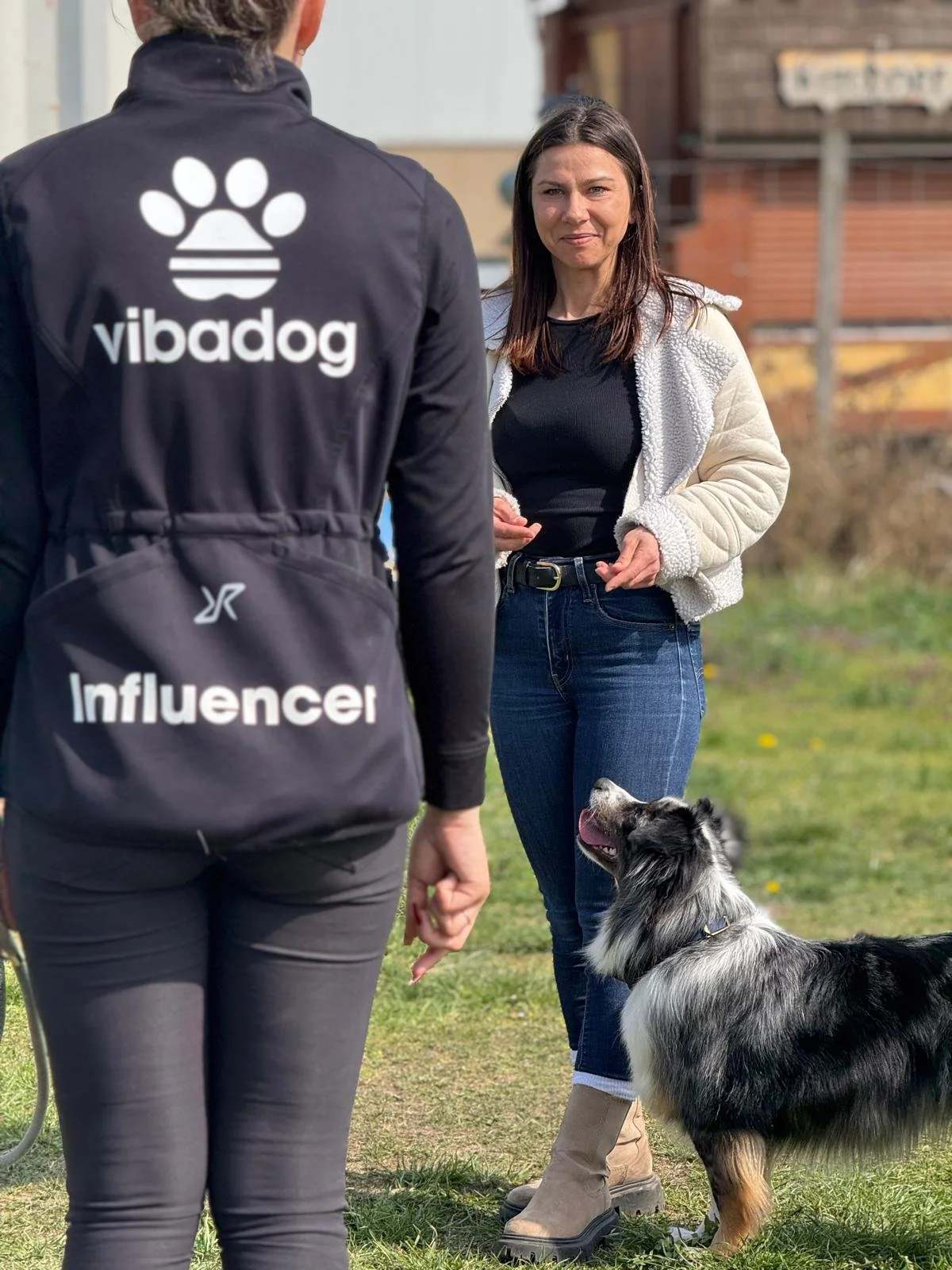 Zwei Frauen sprechen im Freien, eine mit einem Hund. Die Frau auf der linken trägt eine schwarze Jacke mit dem Logo 'vibadog' und der Bezeichnung 'Influencer', die andere trägt eine weiße Jacke und Jeans. Der Hund ist ein Border Collie, der auf den Menschen schaut.