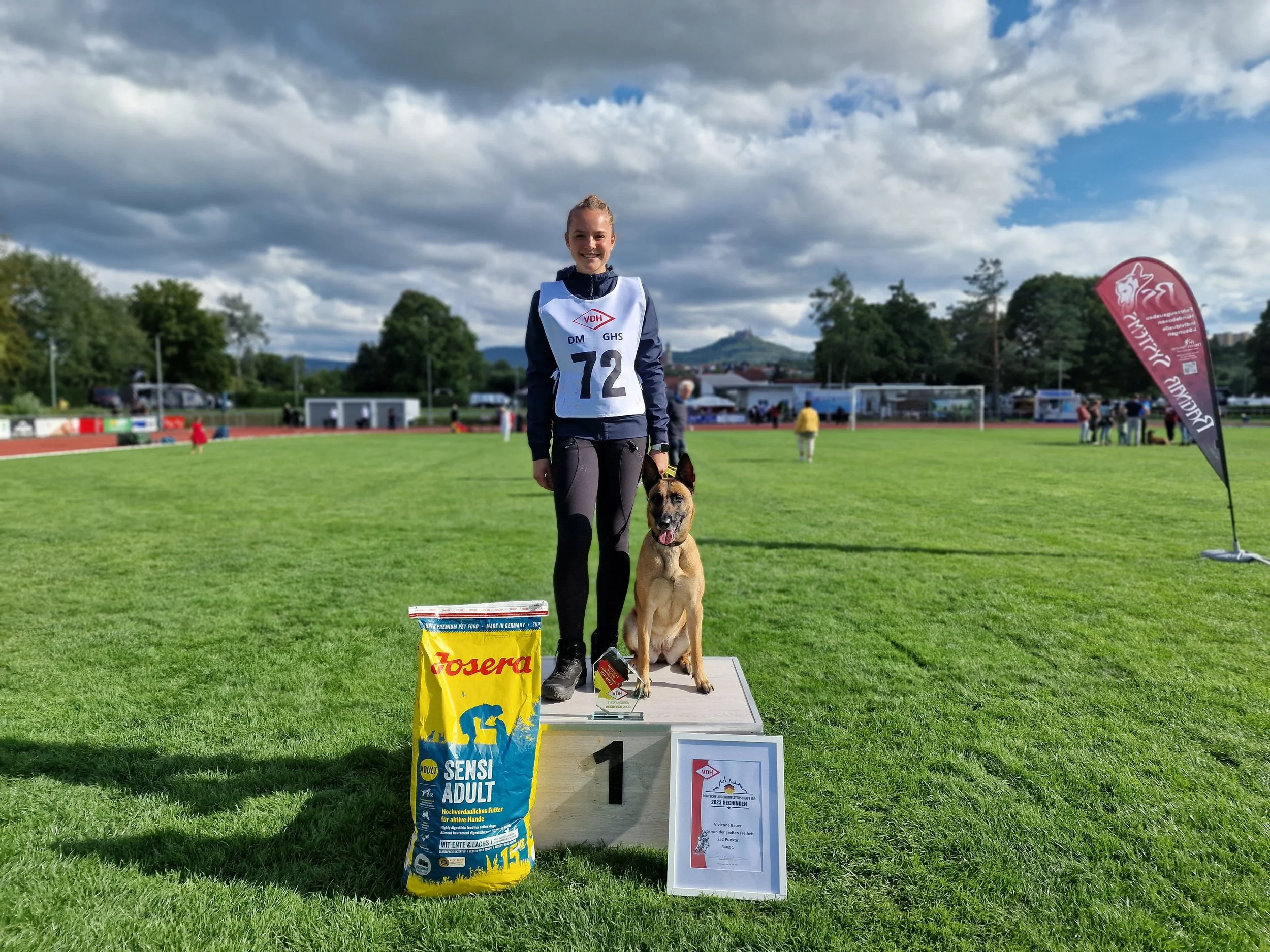 Eine junge Frau steht auf einem Siegerpodest mit einem Hund beim Hundesportwettbewerb. Sie trägt einen Wettkampfanzug mit der Nummer 72. Im Hintergrund sind mehr Menschen, Zelte und ein Sportfeld sichtbar. Das Wetter ist bewölkt.