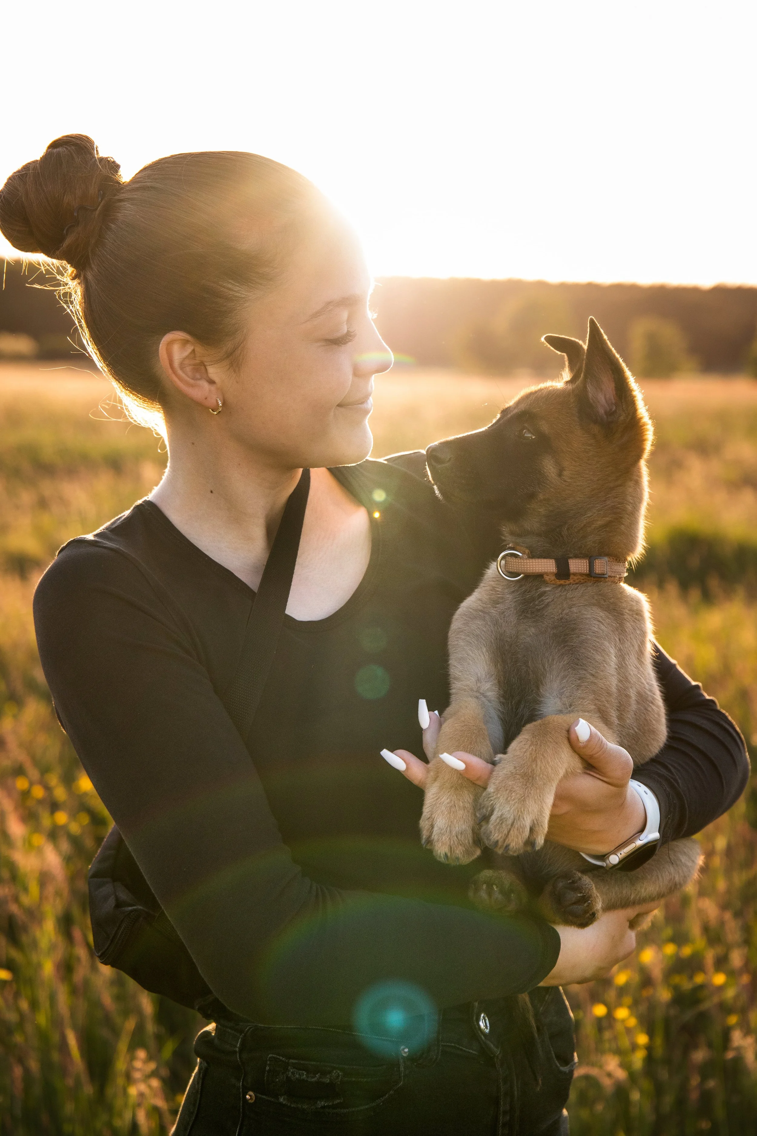 Eine junge Frau hält einen Welpen im Sonnenuntergang, umgeben von einer offenen Wiese und blühenden Pflanzen.