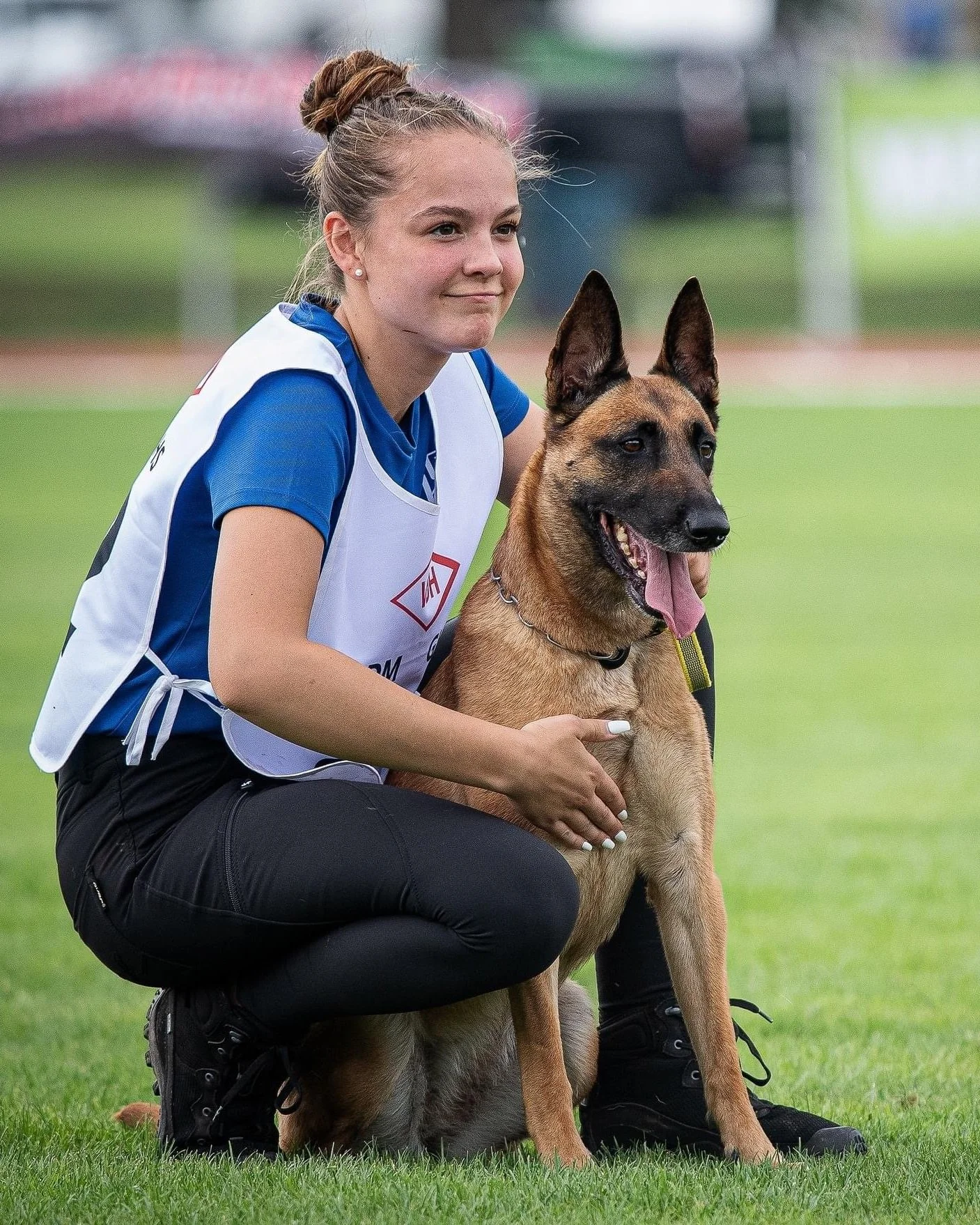 Junge Frau in Sportkleidung mit Polizei-Weste und Schweizer Flagge auf dem Shirt, die mit einem Deutschen Schäferhund auf einem Sportplatz posiert.
