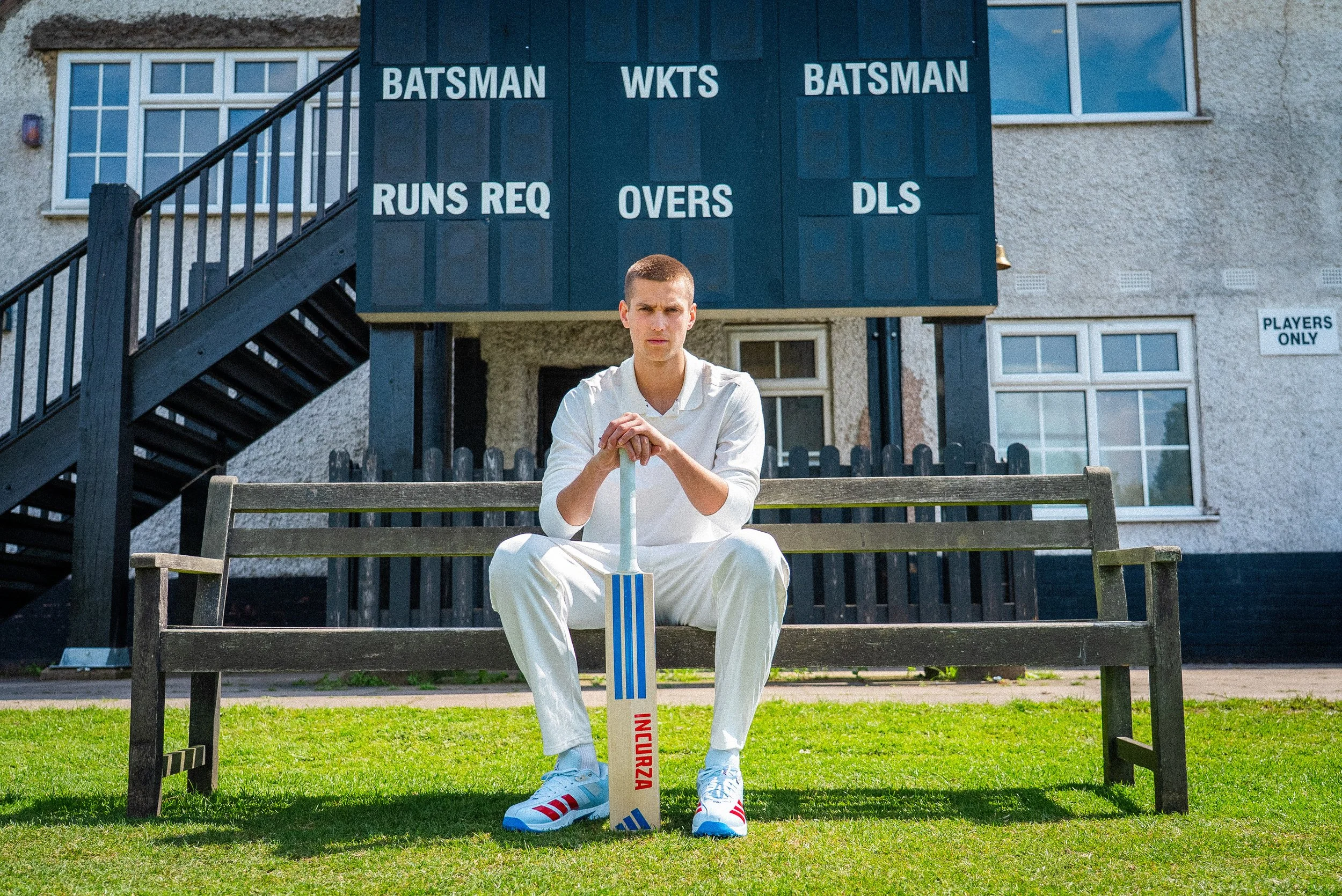 Cricketer in white uniform sitting on a bench with a bat in front of a cricket scoreboard
