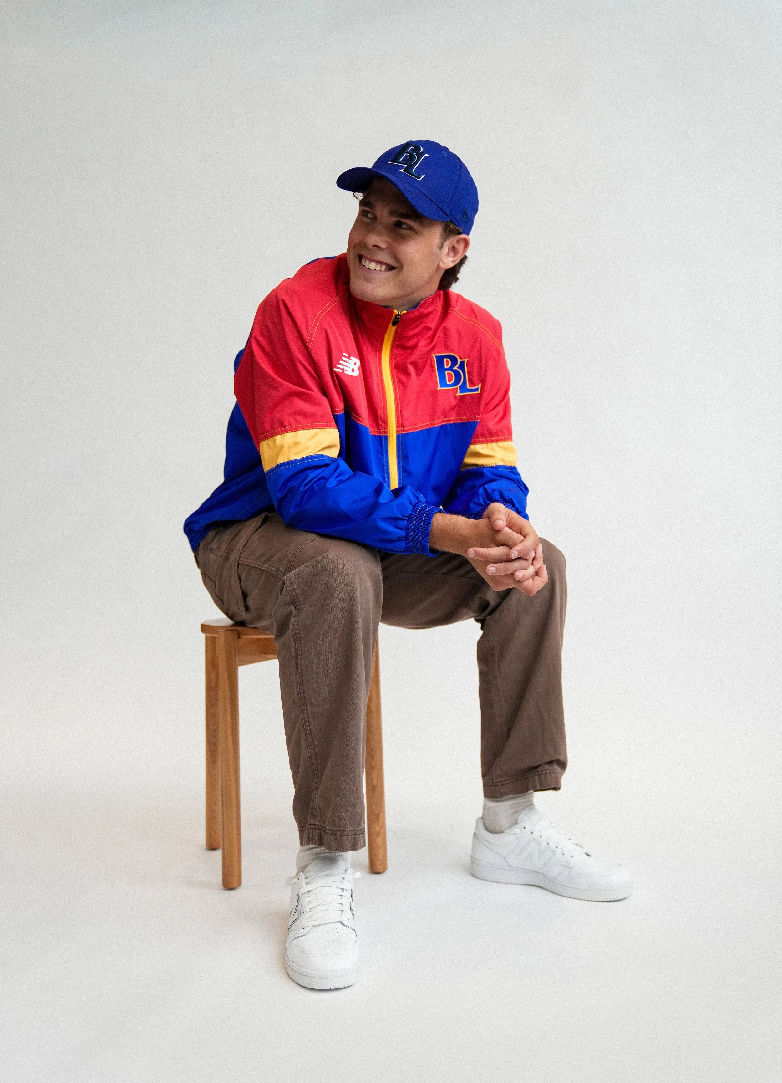 Smiling man in a blue BL baseball cap and colorful red, blue, and yellow jacket sitting on a wooden stool against a white background
