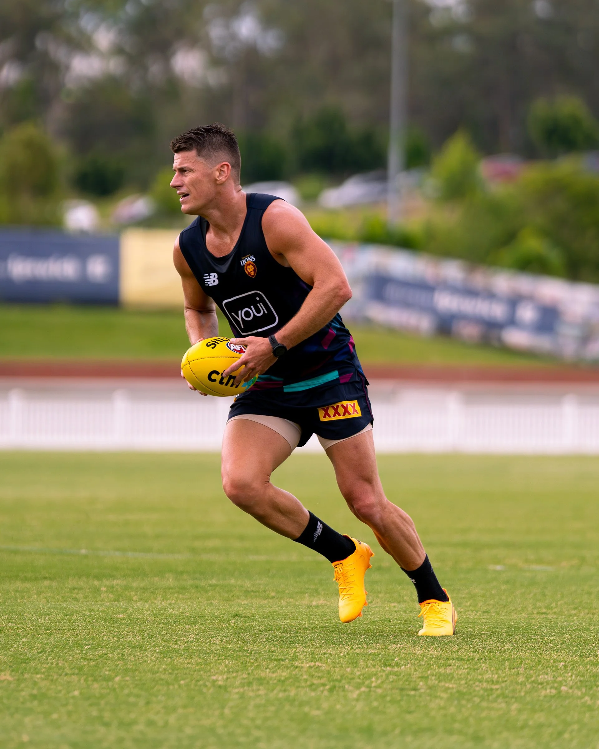Australian rules football player in a Brisbane Lions uniform running with a yellow football on a grass field