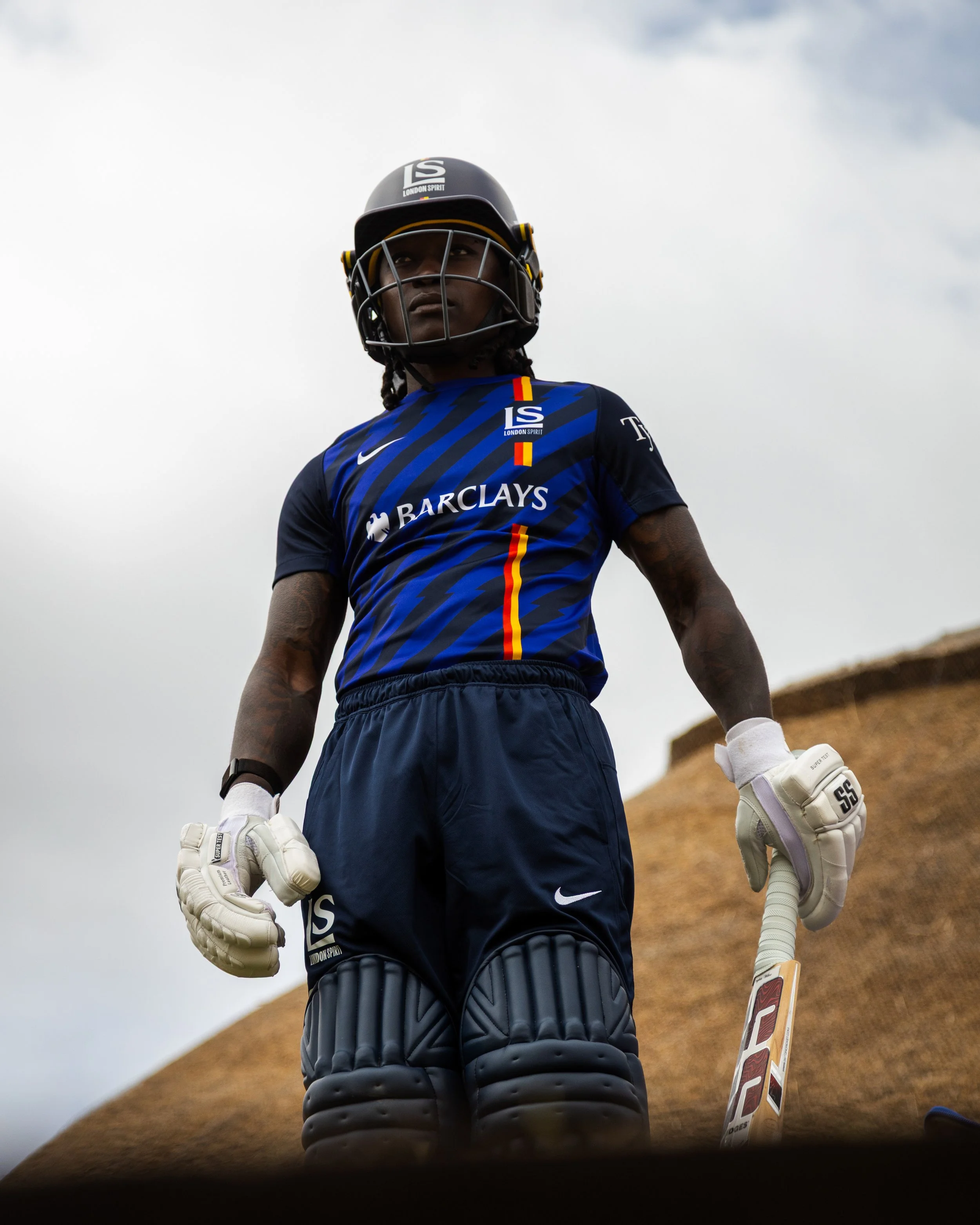 London Spirit cricket player wearing a helmet, blue jersey, leg pads, and batting gloves while holding a cricket bat.