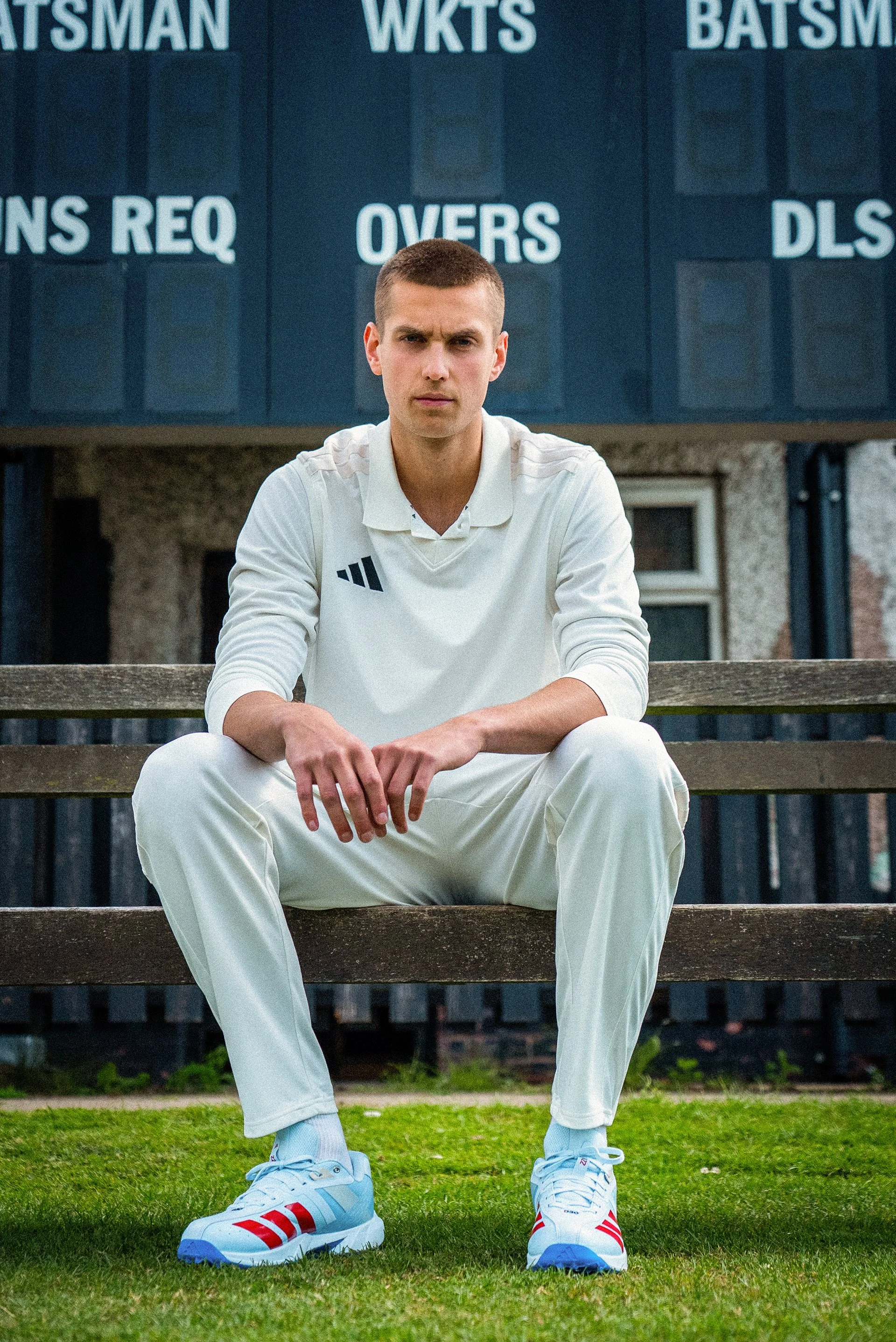 Man in a white Adidas outfit sitting on outdoor bleachers, wearing white and red Adidas sneakers