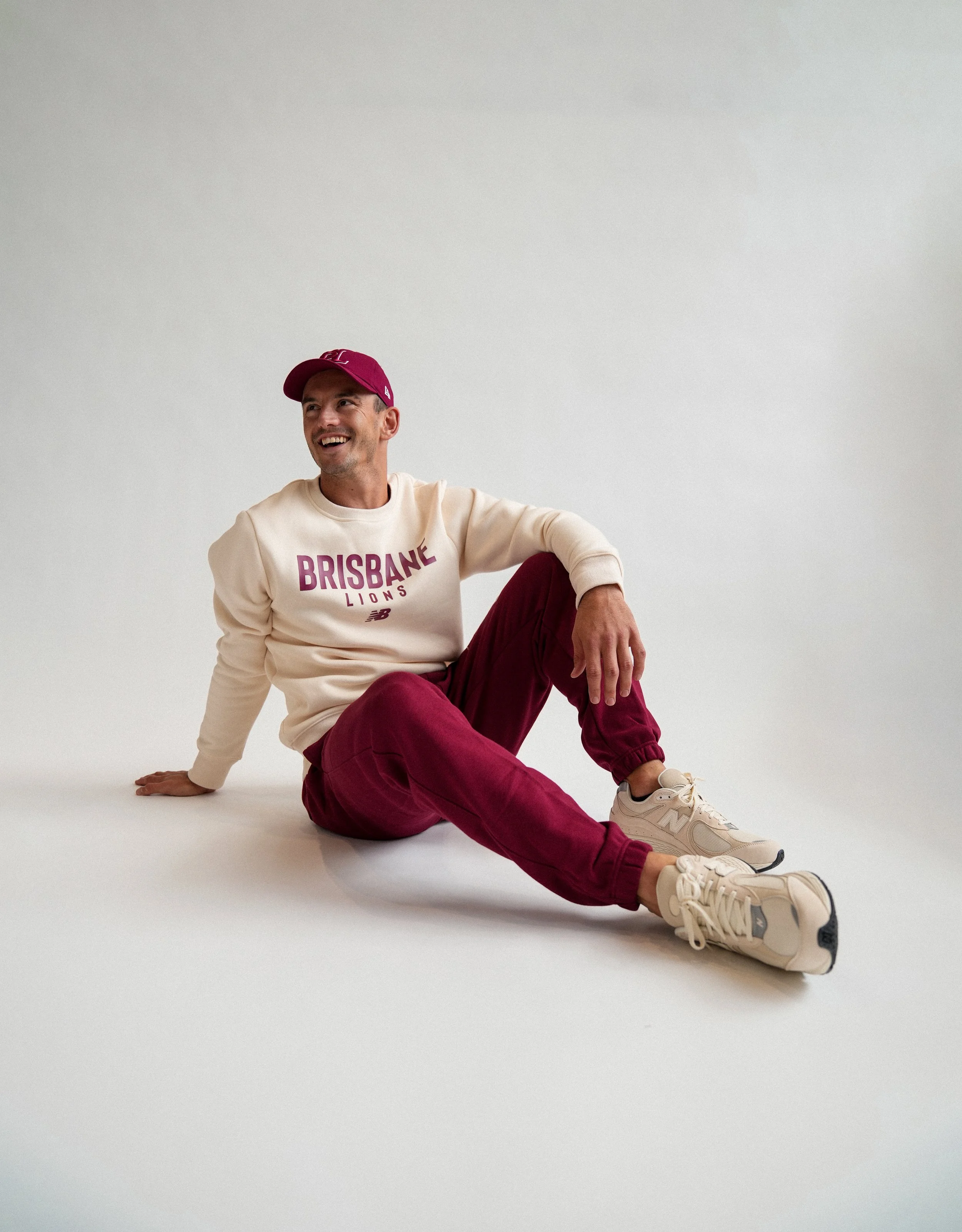 Smiling man wearing Brisbane Lions sweatshirt, burgundy pants, cap, and New Balance sneakers seated on a white studio floor