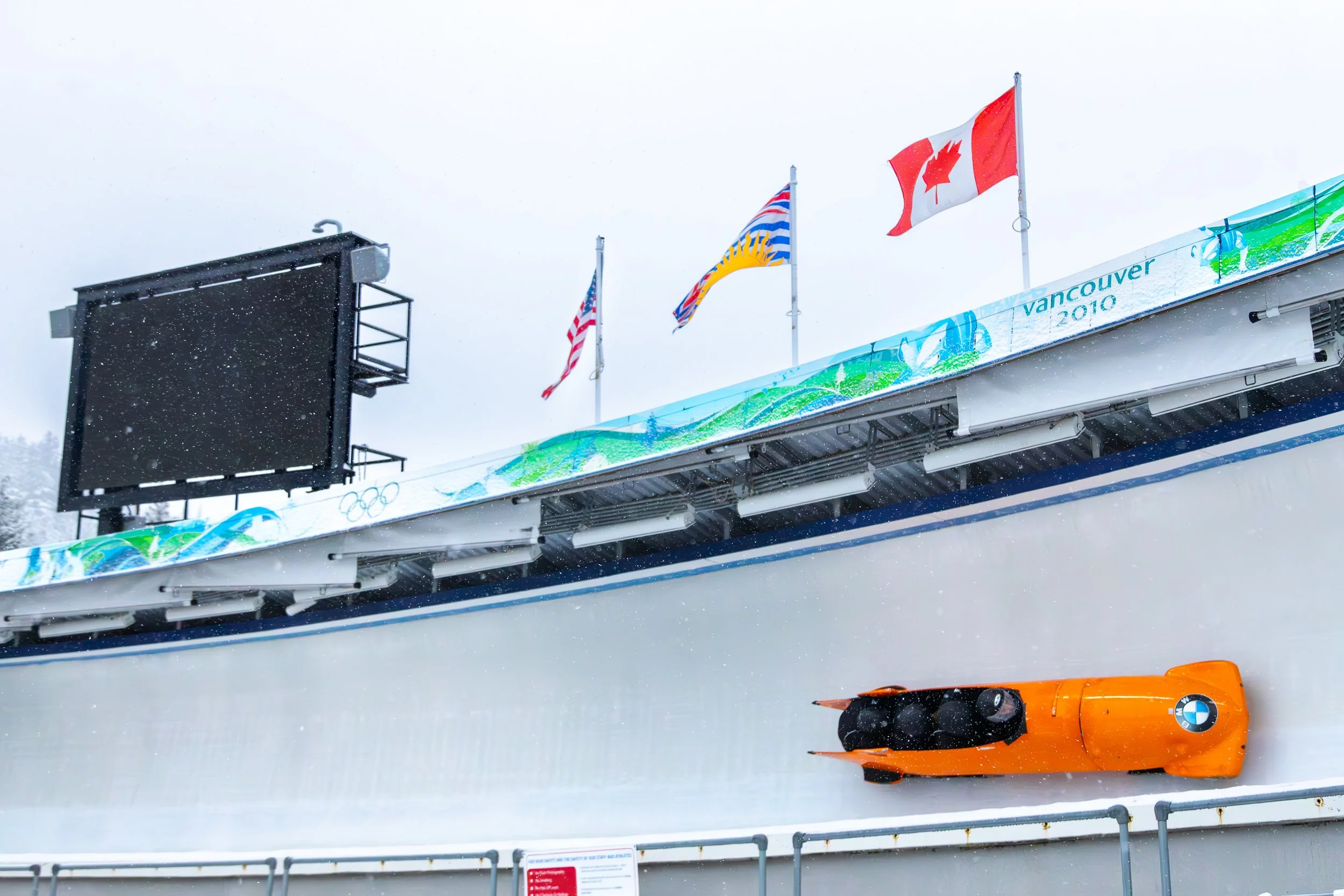 Orange BMW bobsled on a snowy track at the Vancouver 2010 Winter Olympics, with Canadian and international flags flying above.