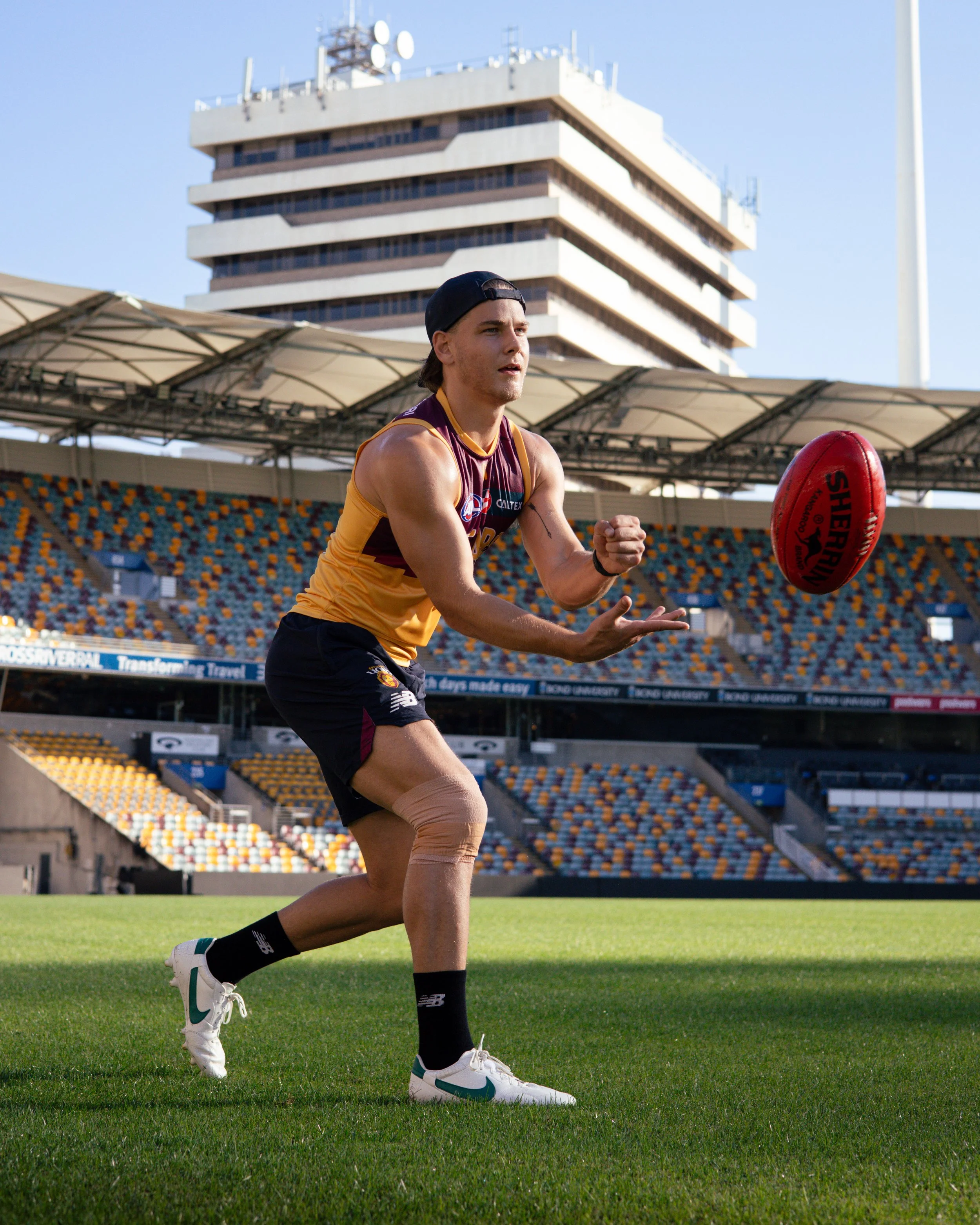 Australian rules football player in a maroon-and-gold uniform reaching for a red football on a stadium field