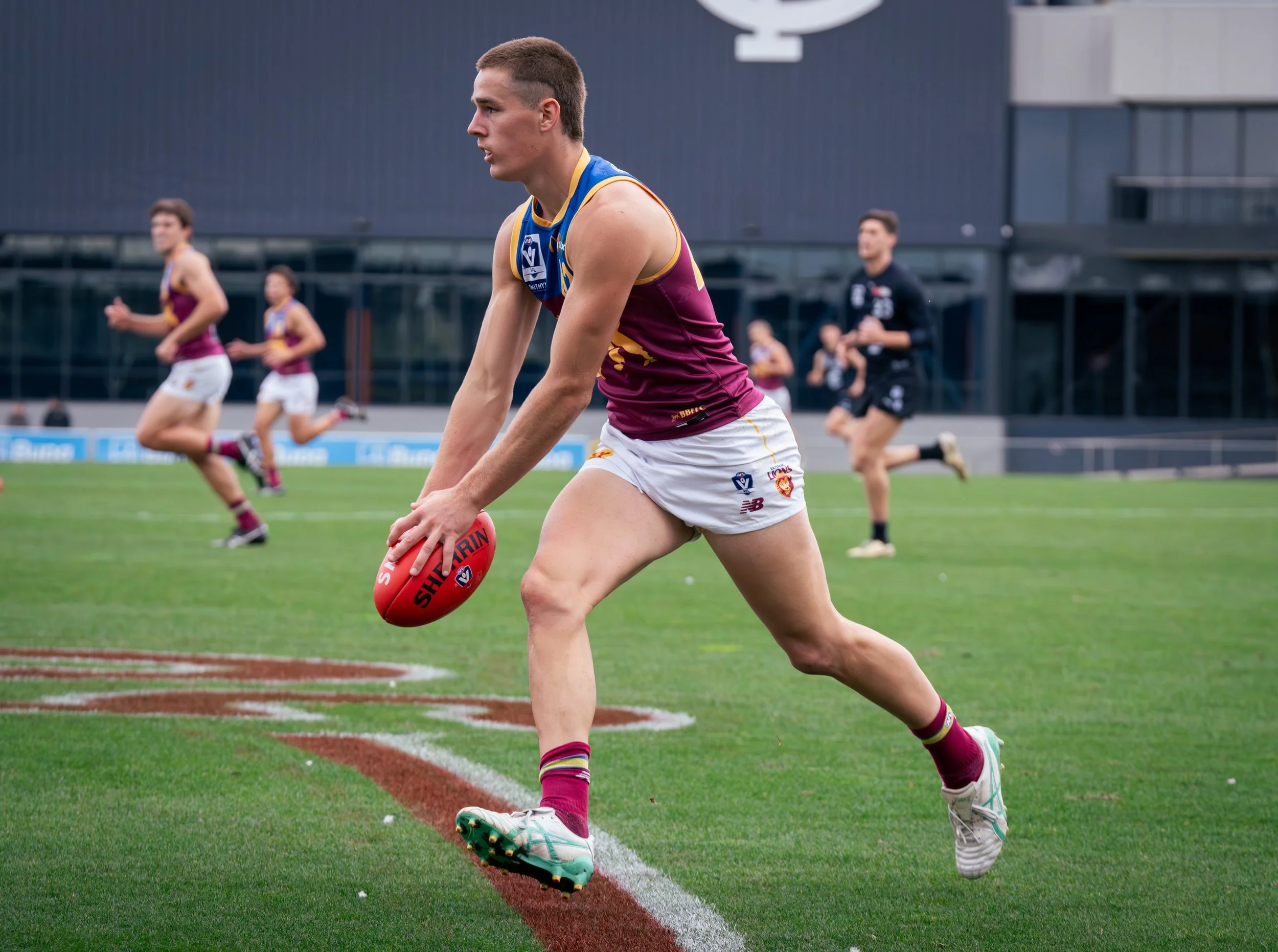 Australian rules football player in a maroon, blue, and gold uniform running with a red football during practice