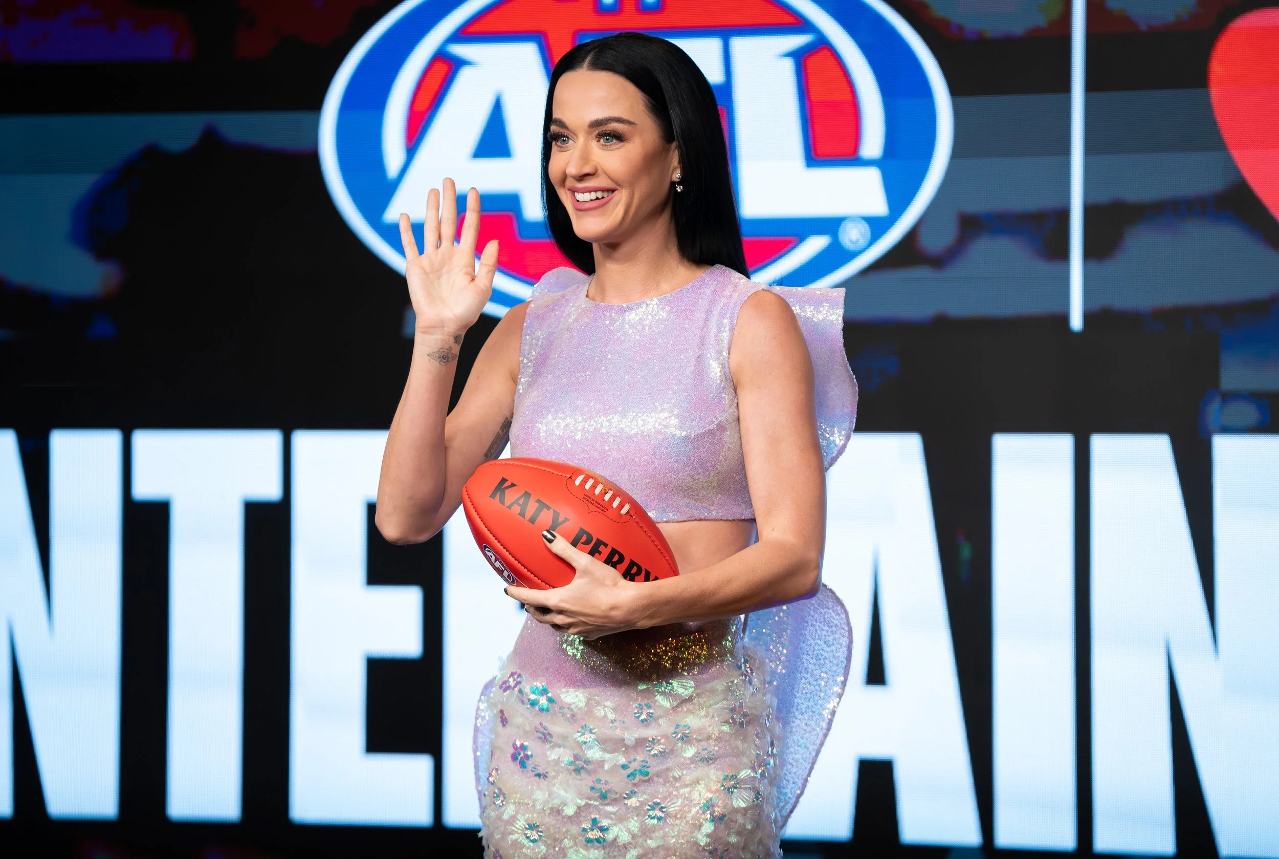 Katy Perry waves while holding a football onstage in front of an American Football League logo.