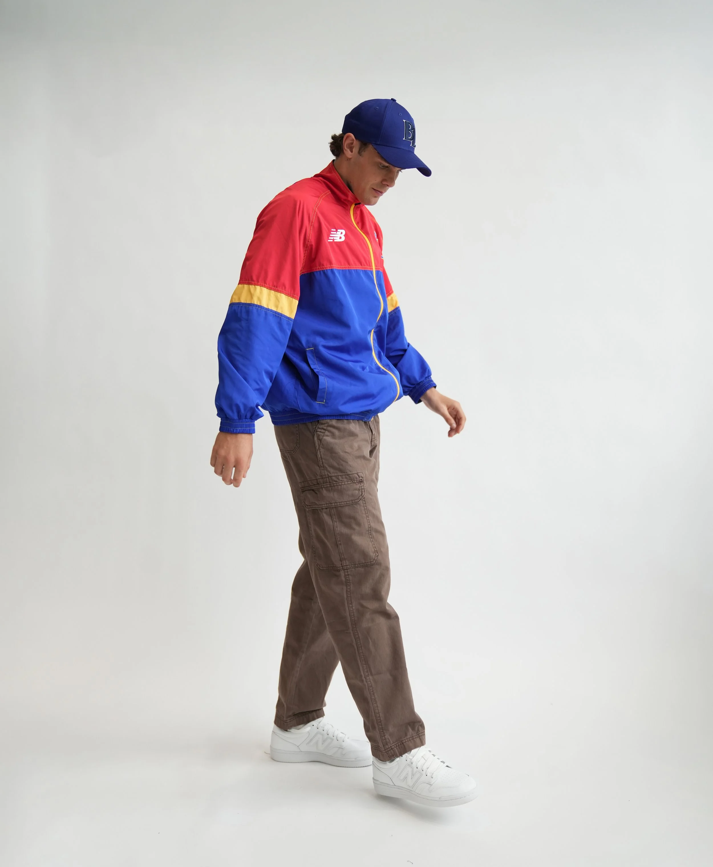 Man wearing a blue, red, and yellow New Balance windbreaker, blue cap, cargo pants, and white sneakers