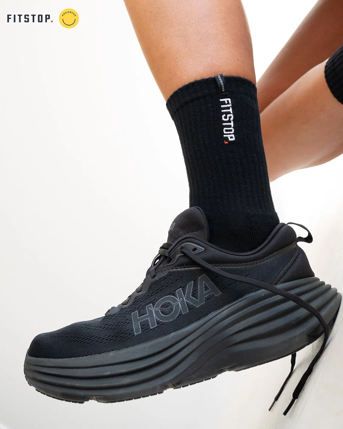 Close-up of a black HOKA sneaker with thick sole and black FITS socks on a person’s foot