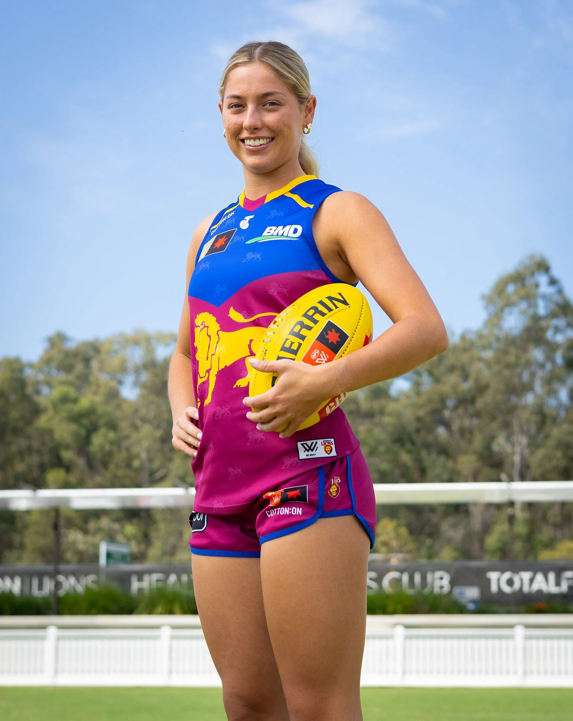 Smiling female Australian rules football player in a Brisbane Lions uniform holding a yellow football outdoors