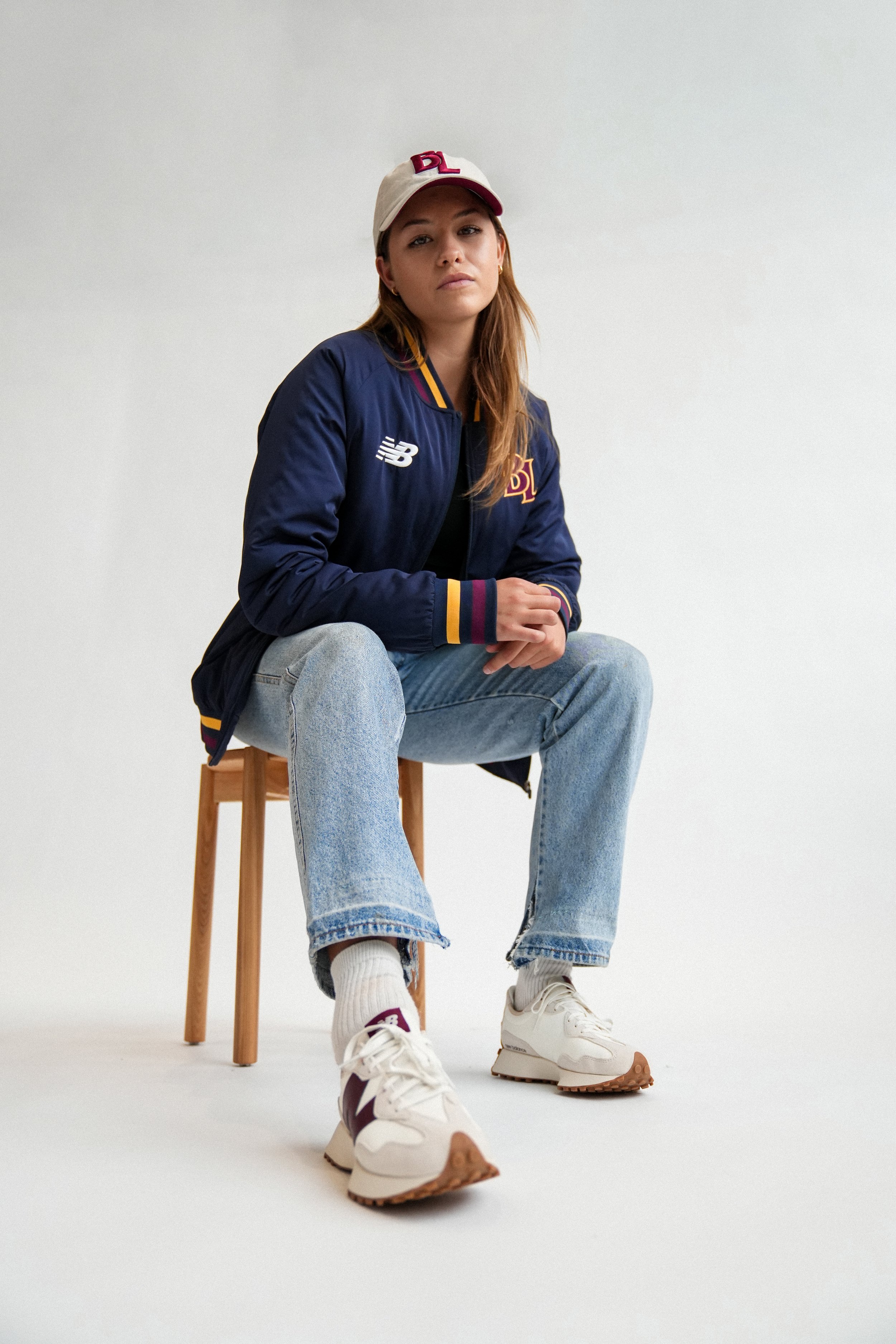 Woman wearing a Boston Red Sox jacket, cap, jeans, and New Balance sneakers while sitting on a wooden stool.