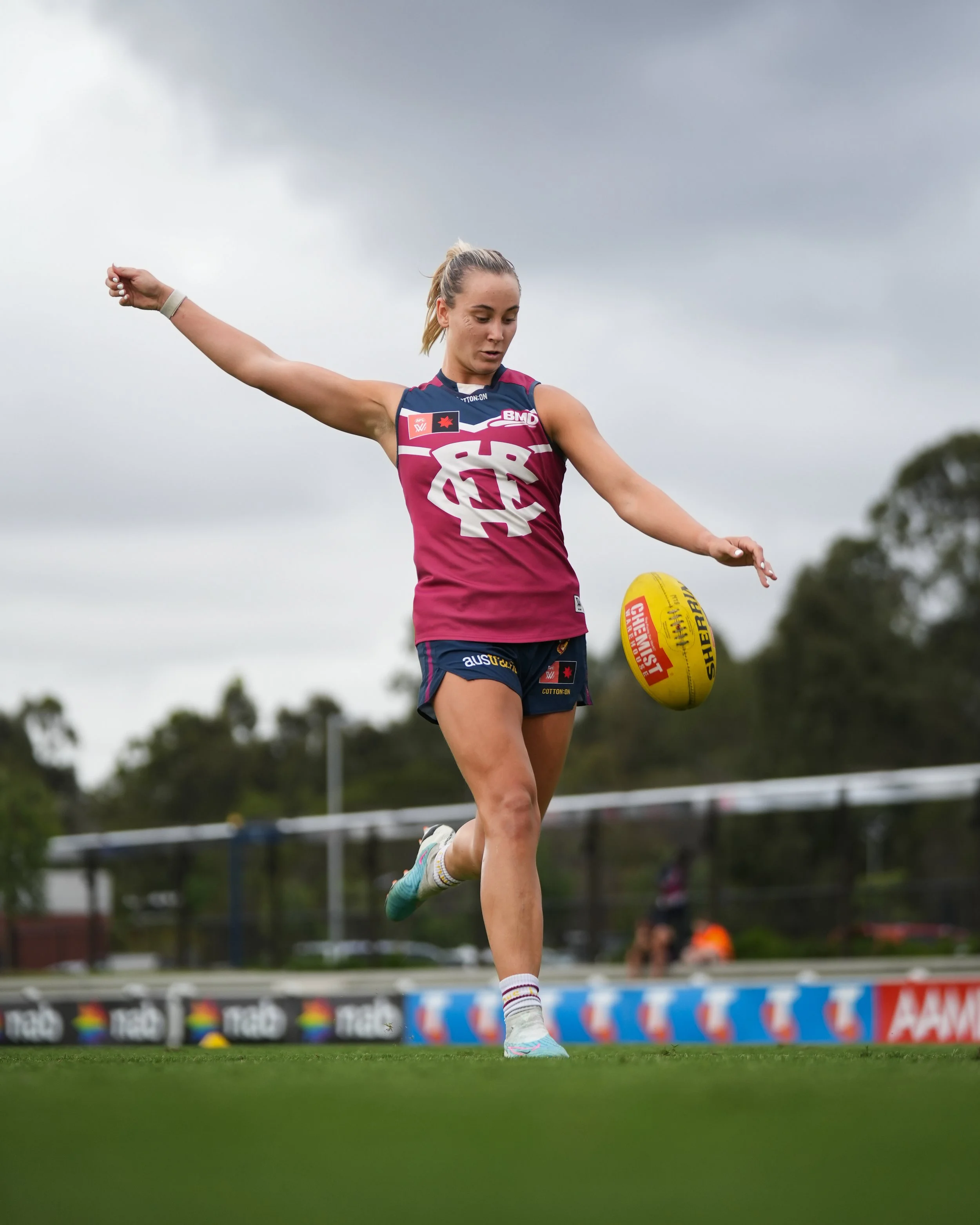 Female Australian rules football player in a magenta uniform kicking a yellow football on a grassy field