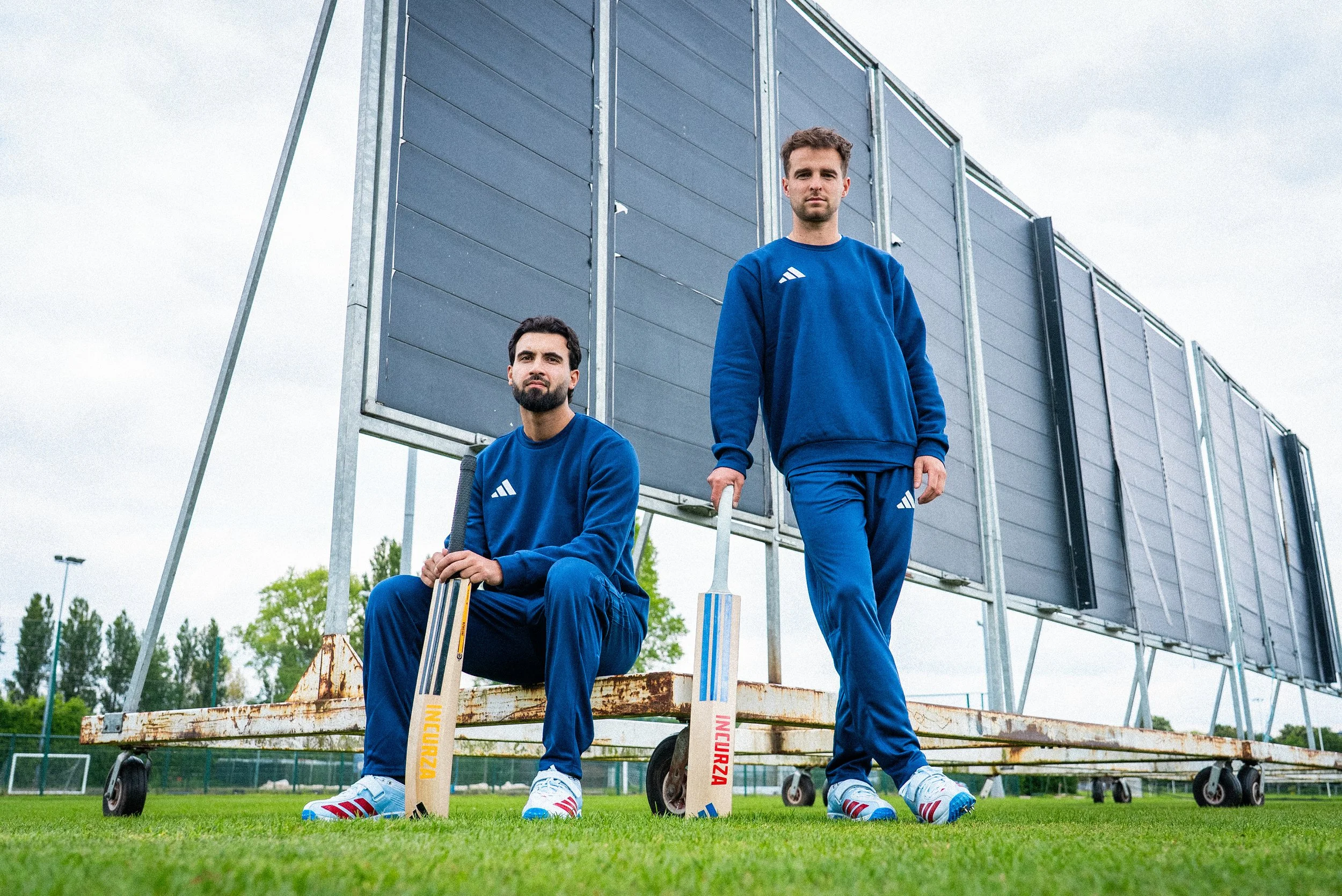Two men in blue Adidas cricket uniforms holding cricket bats on a grassy field