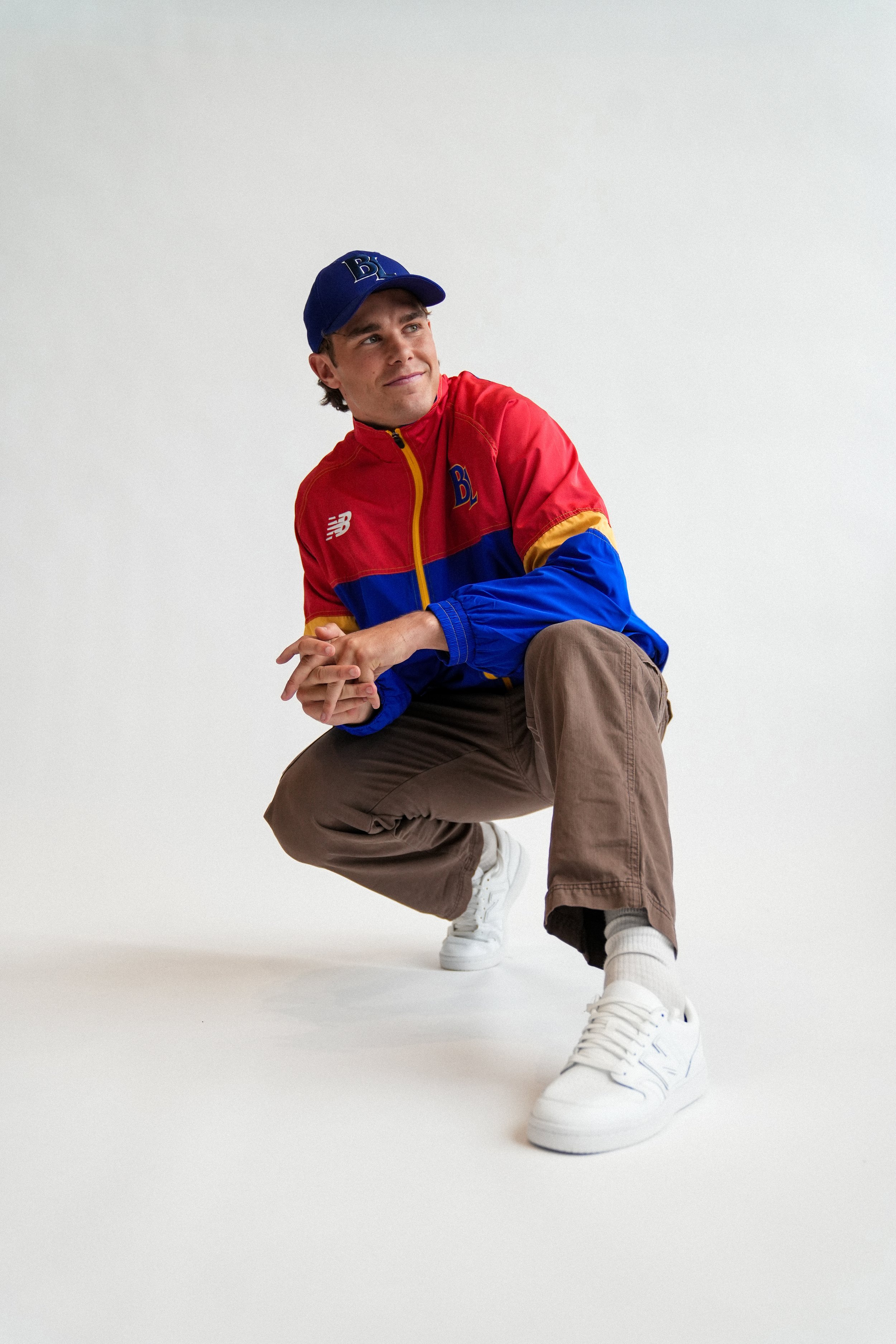 Young man wearing a blue baseball cap and colorful red-and-blue jacket, seated against a white background