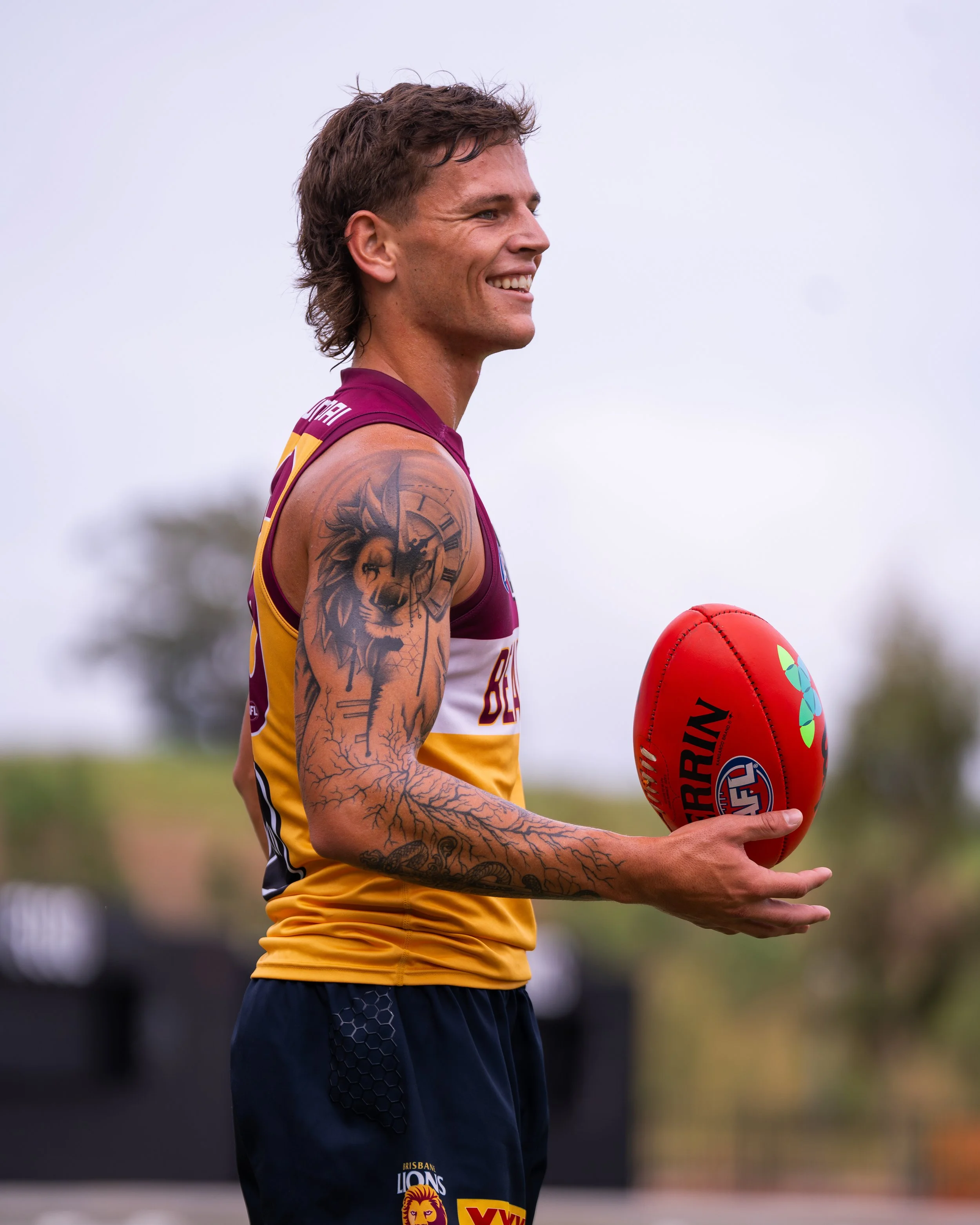 Tattooed Brisbane Lions Australian rules football player smiling while holding a red Sherrin football.