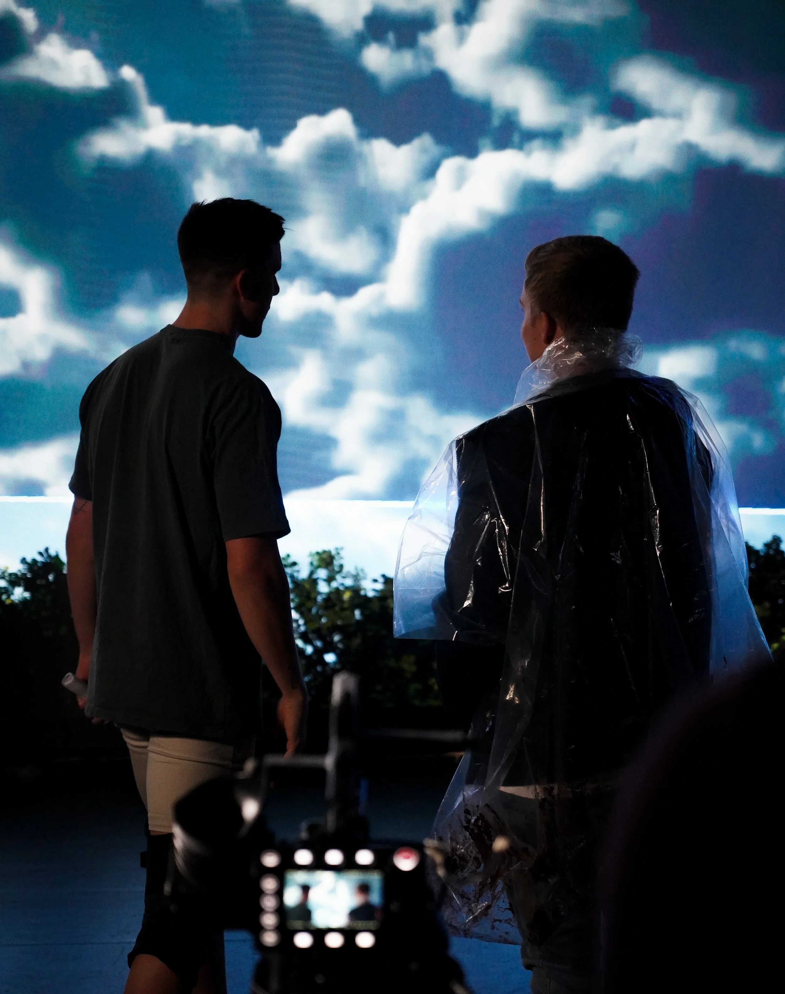 Two people in silhouette watch a large projected cloudscape while one wears a clear rain poncho and a camera is visible in the foreground.
