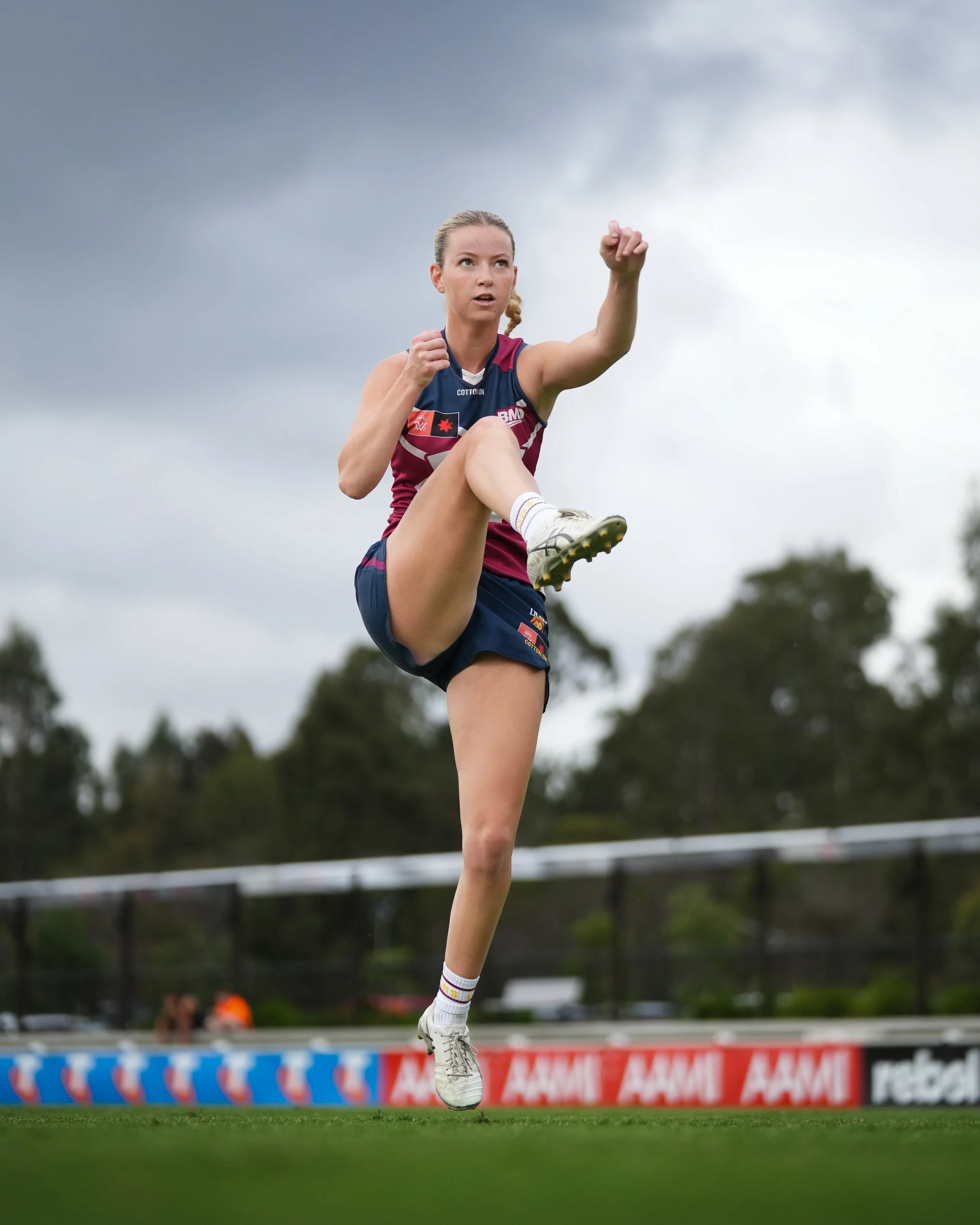 Female Australian rules football player leaping to kick a ball on a field