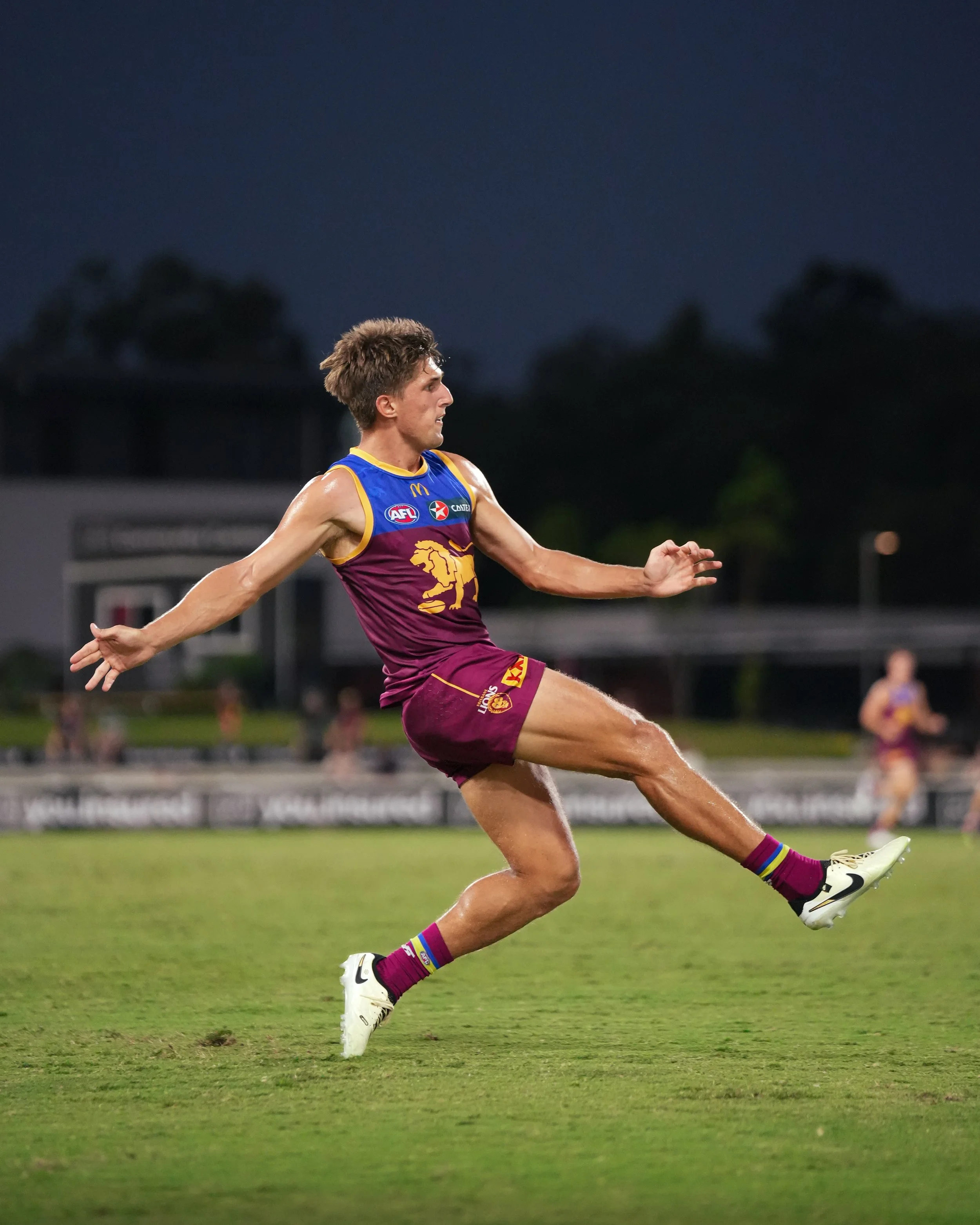 Brisbane Lions AFL player running across the field with arms outstretched during a nighttime match.