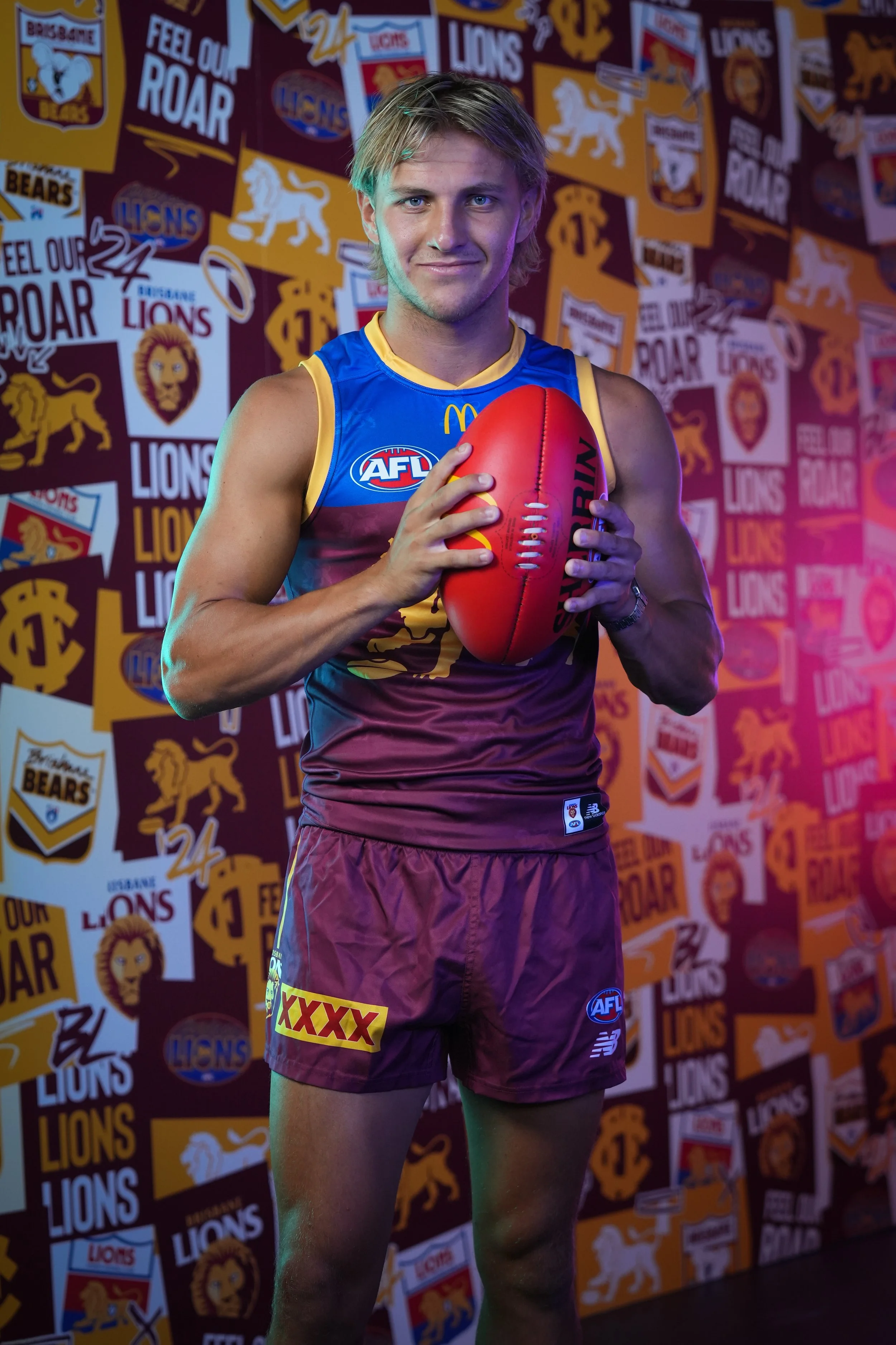Brisbane Lions AFL player holding a red football in front of a team-logo backdrop