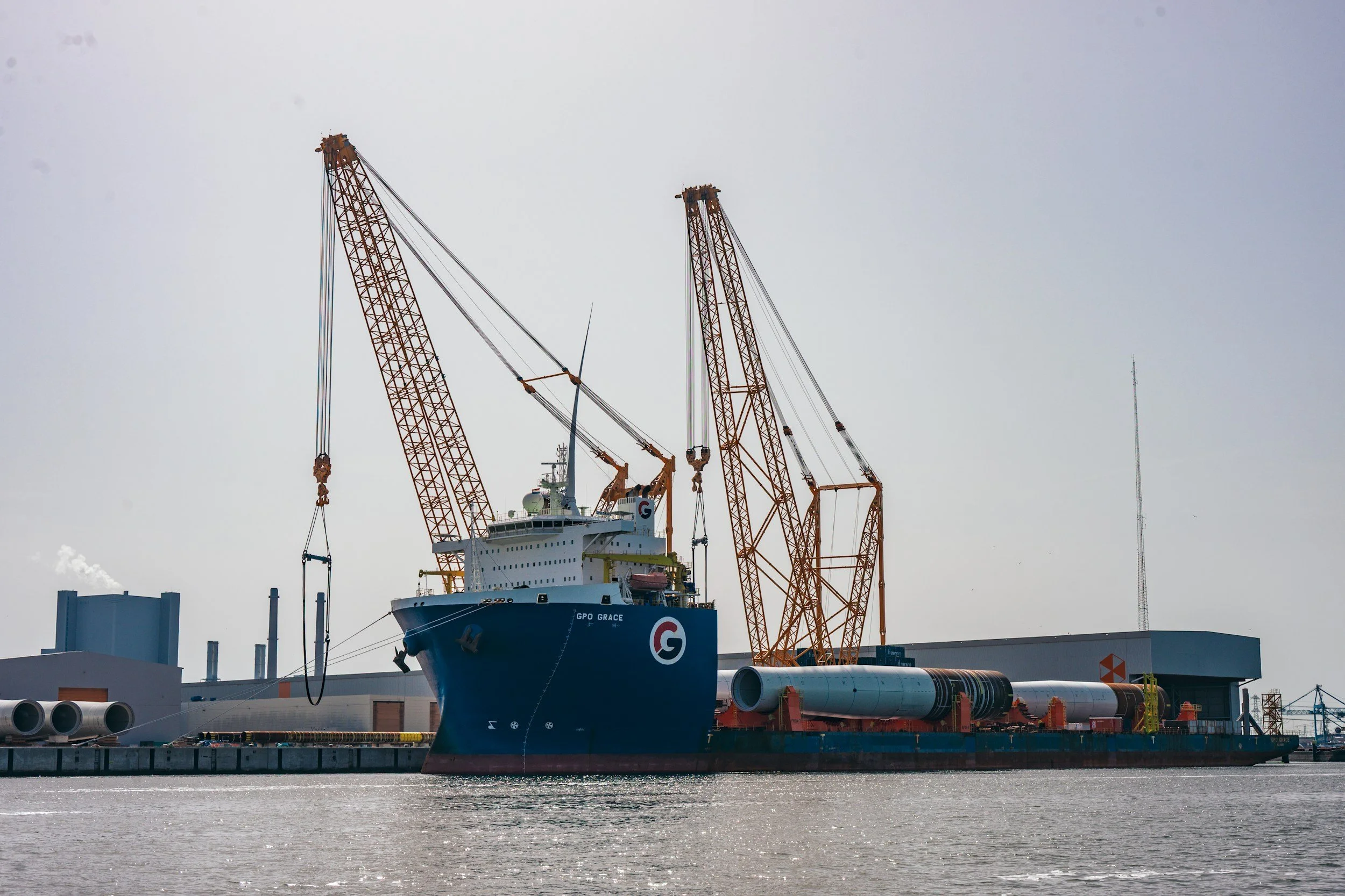 Heavy-lift cargo ship GPO Grace docked at an industrial port with large cranes and cylindrical steel cargo