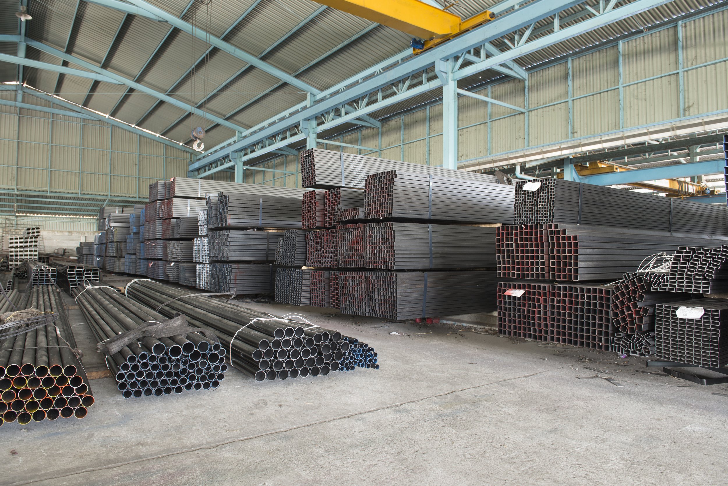 Industrial steel warehouse filled with stacked metal pipes, hollow sections, and overhead cranes