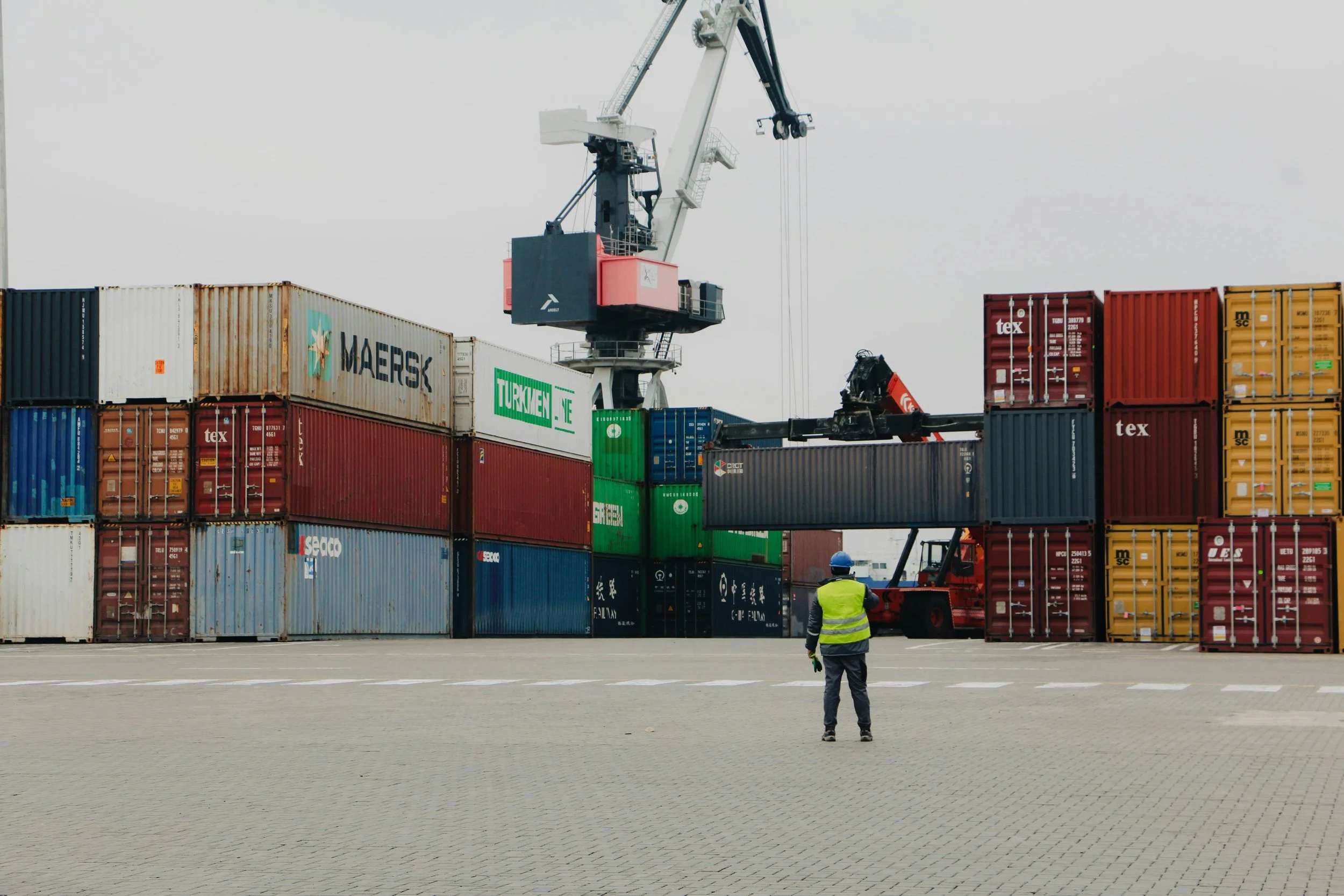 Port crane loading shipping containers at a busy cargo terminal, with a worker in a high-visibility vest.