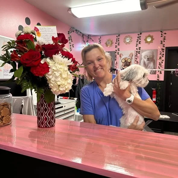 Woman holding a small dog behind a pink counter beside a vase of red and white flowers