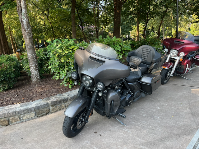 Black touring motorcycle parked on a driveway with a red touring motorcycle behind it, surrounded by landscaped trees and shrubs