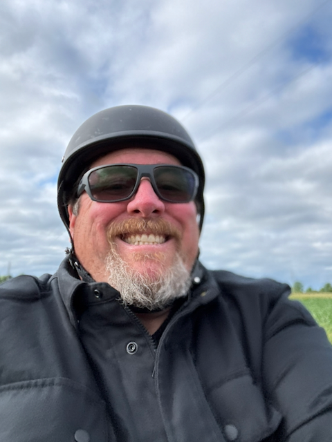 Smiling man wearing a black motorcycle helmet, sunglasses, and jacket outdoors under a cloudy sky