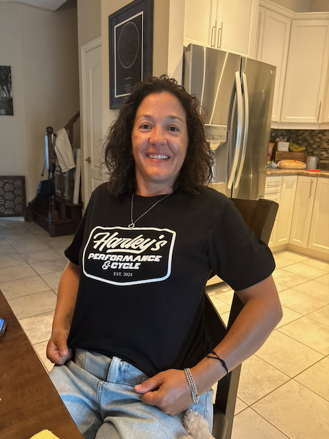 Smiling woman wearing a black “Honey’s Performance Cycle” T-shirt seated in a bright home kitchen