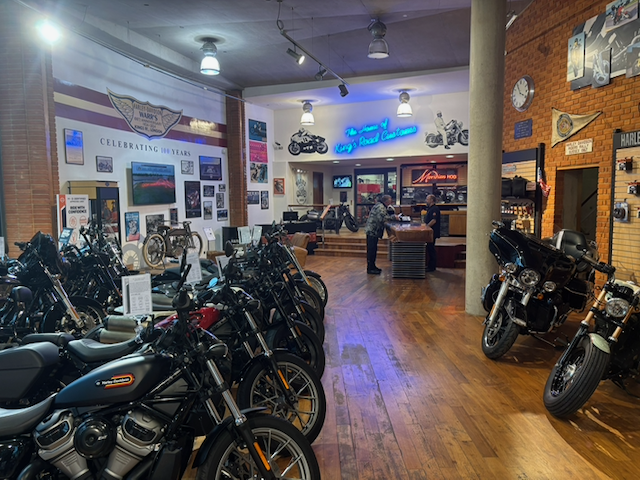 Interior of a Harley-Davidson motorcycle dealership displaying rows of motorcycles, vintage memorabilia, branded signs, and a customer service counter.