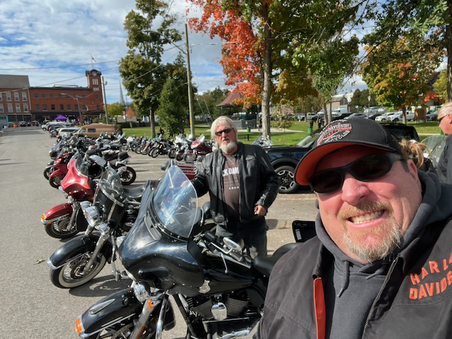 Harley-Davidson riders pose beside a lineup of motorcycles on a sunny autumn day.