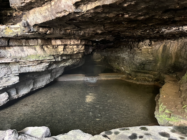 Rocky cave chamber with layered limestone walls and a shallow, clear pool of water