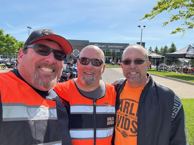 Three smiling Harley-Davidson riders posing outdoors in front of a Harley-Davidson dealership