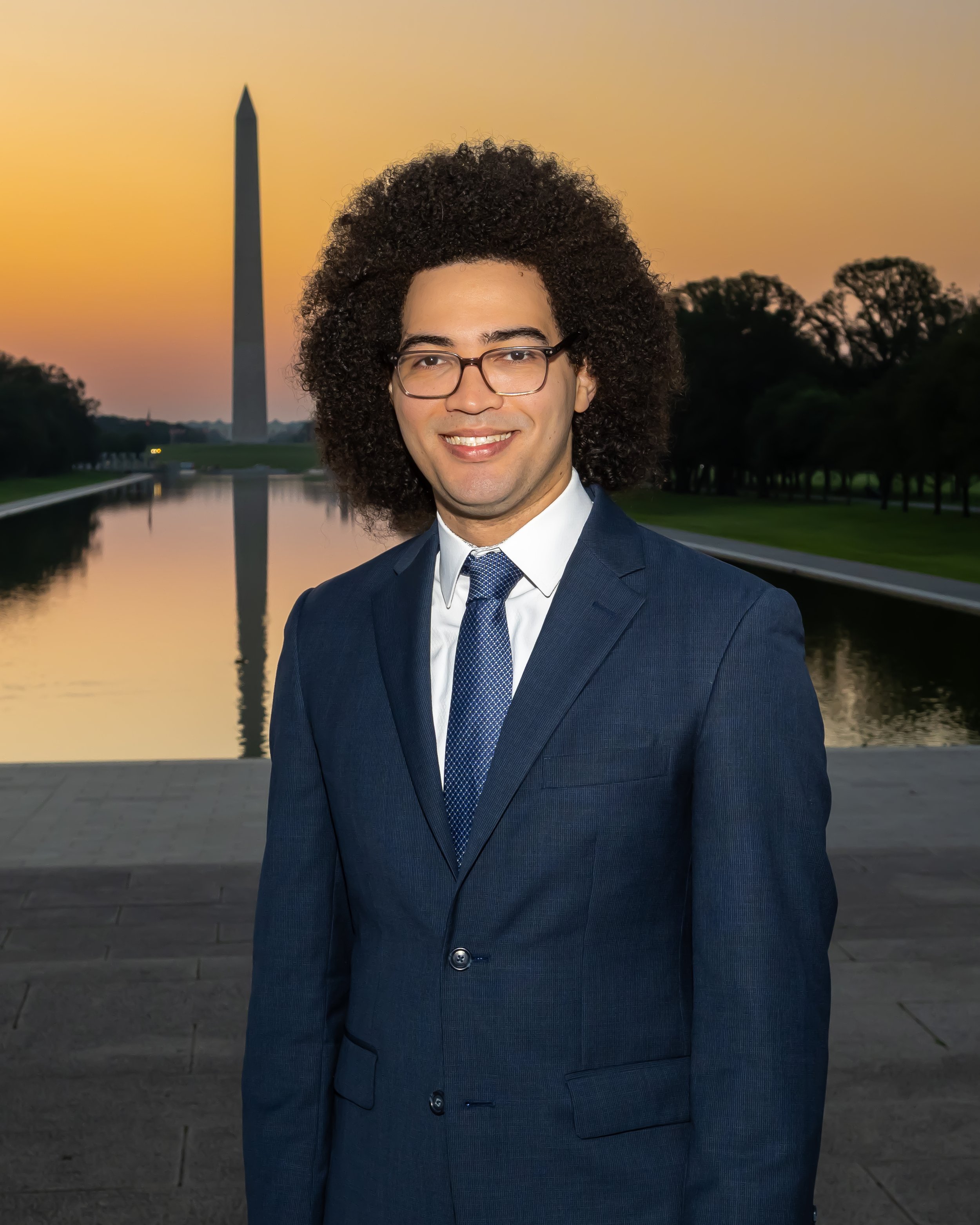 A man with curly hair and glasses smiling, wearing a navy blue suit and tie, standing outdoors at sunset with the Washington Monument and trees reflected in a waterway behind him.