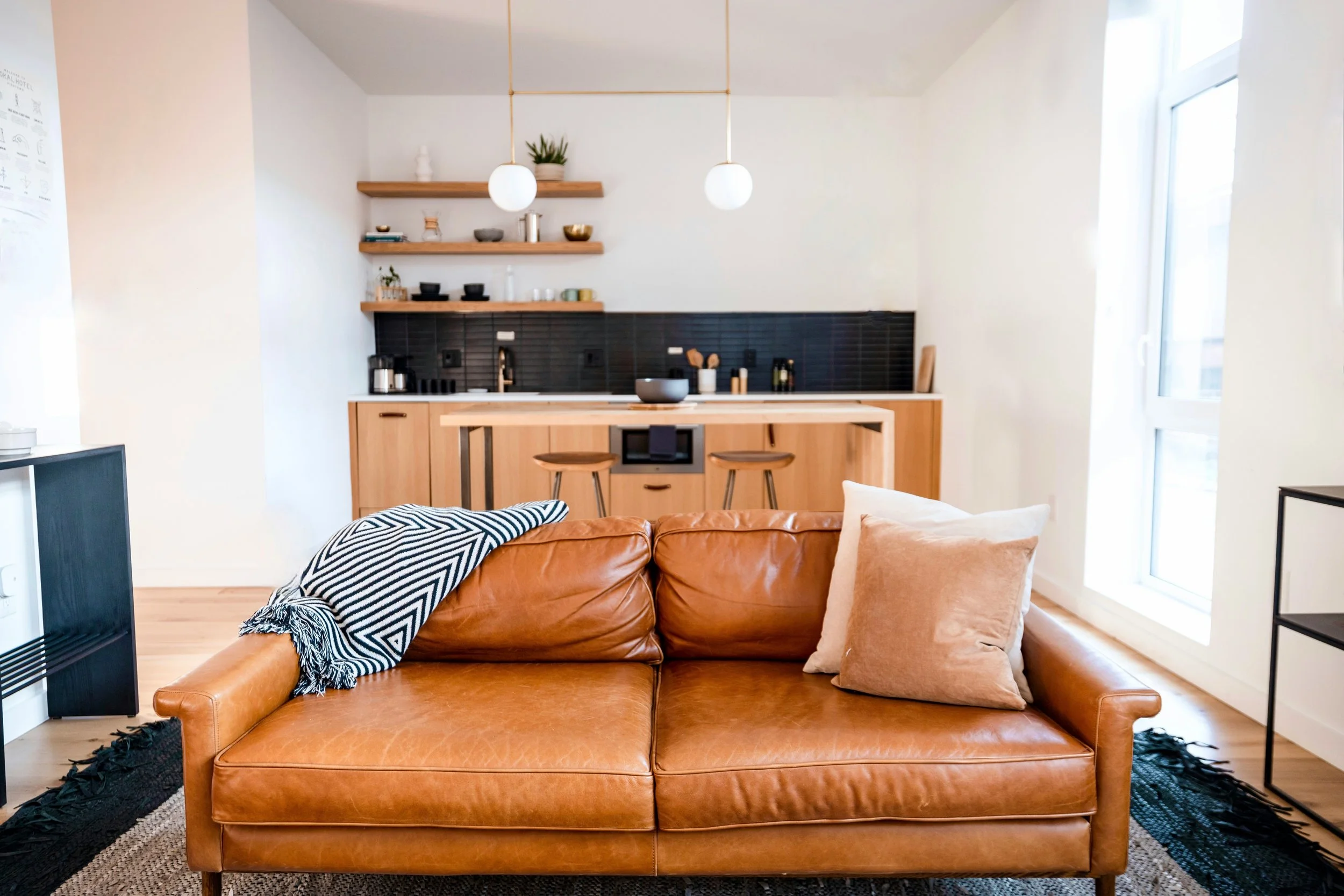 Modern open-plan living room with a caramel leather sofa, patterned throw blanket, pillows, and adjacent kitchen with wood cabinets and black backsplash