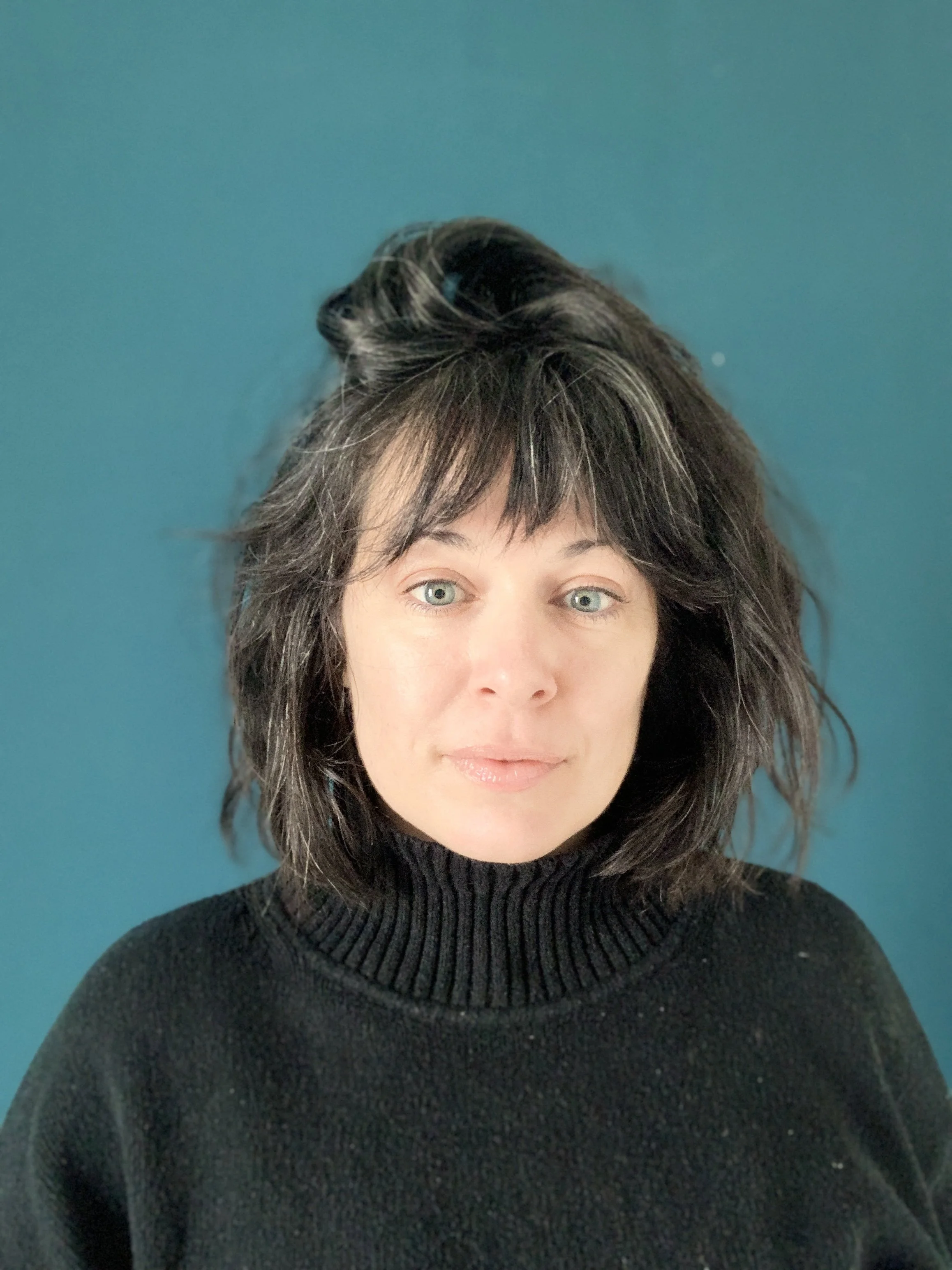 Close-up portrait of a woman with dark hair and bangs wearing a black turtleneck against a teal background