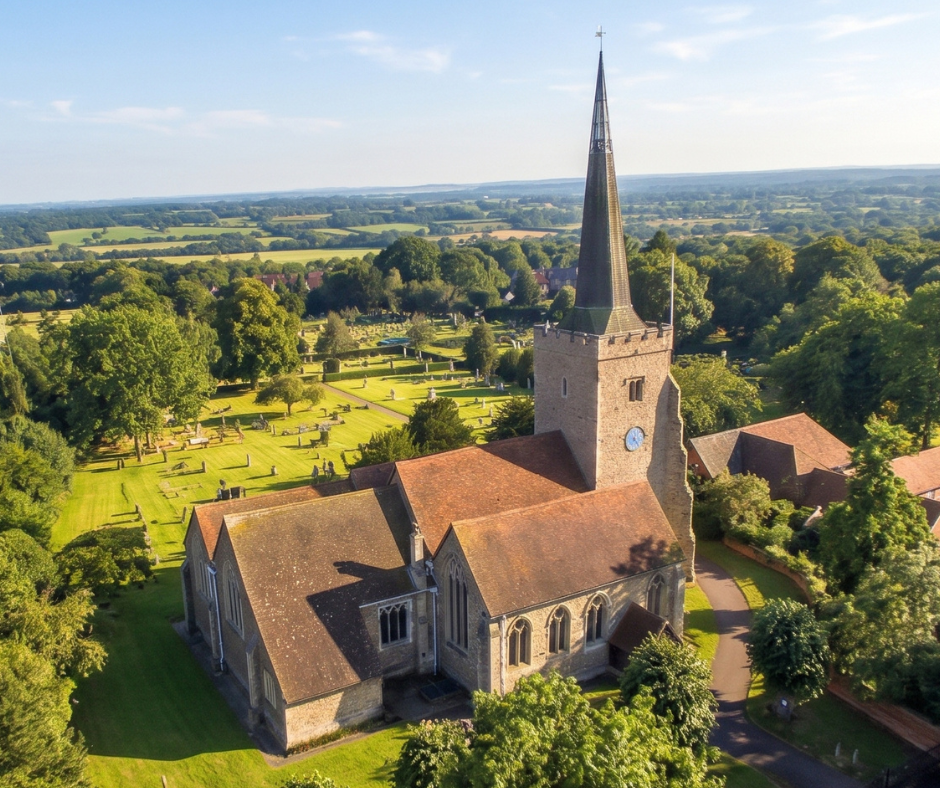 Aerial view of a historic stone church with a tall steeple, surrounded by a churchyard, trees, and rolling English countryside.
