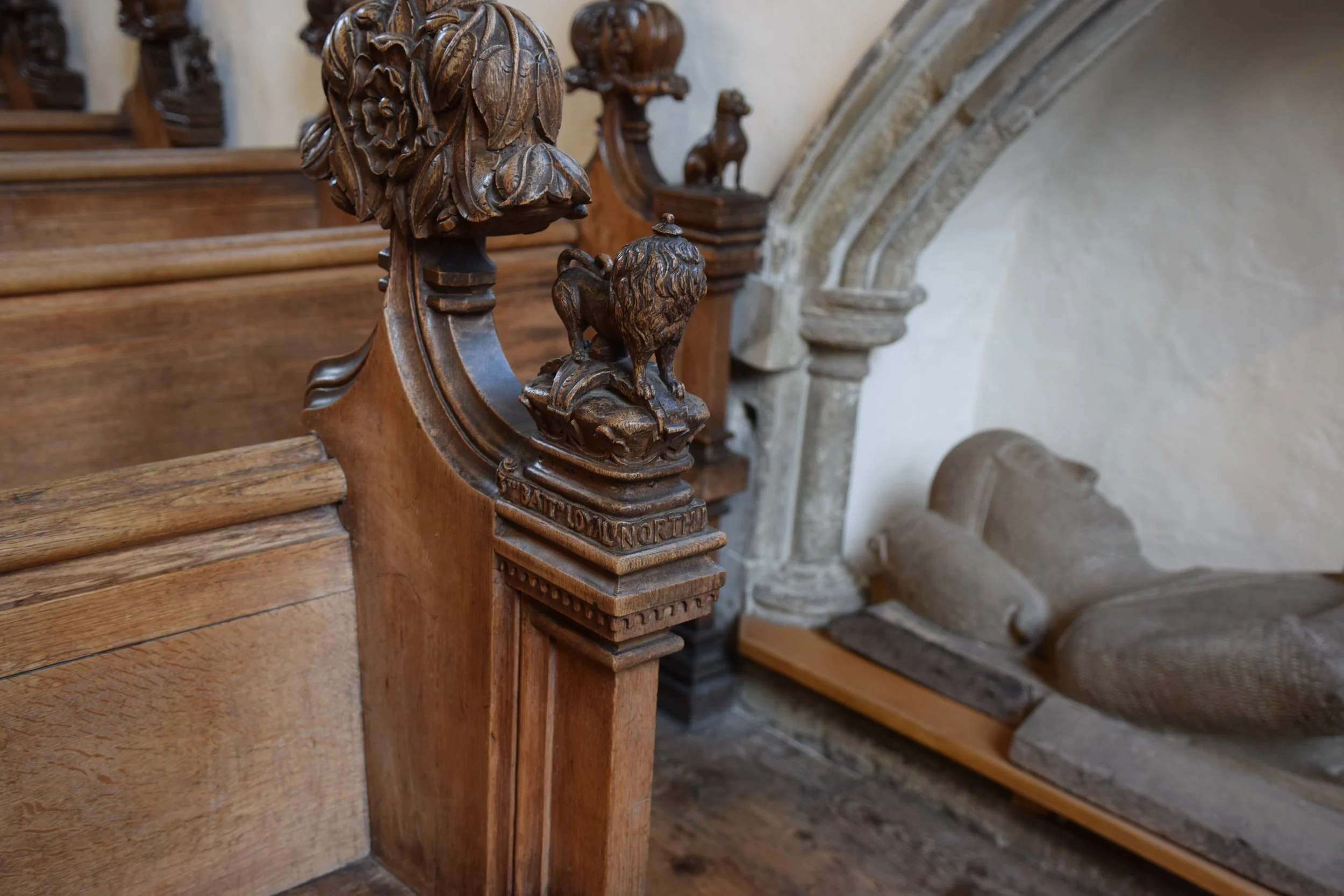 Close-up of an ornate carved wooden church pew end with a lion finial and floral detailing beside a stone arch.