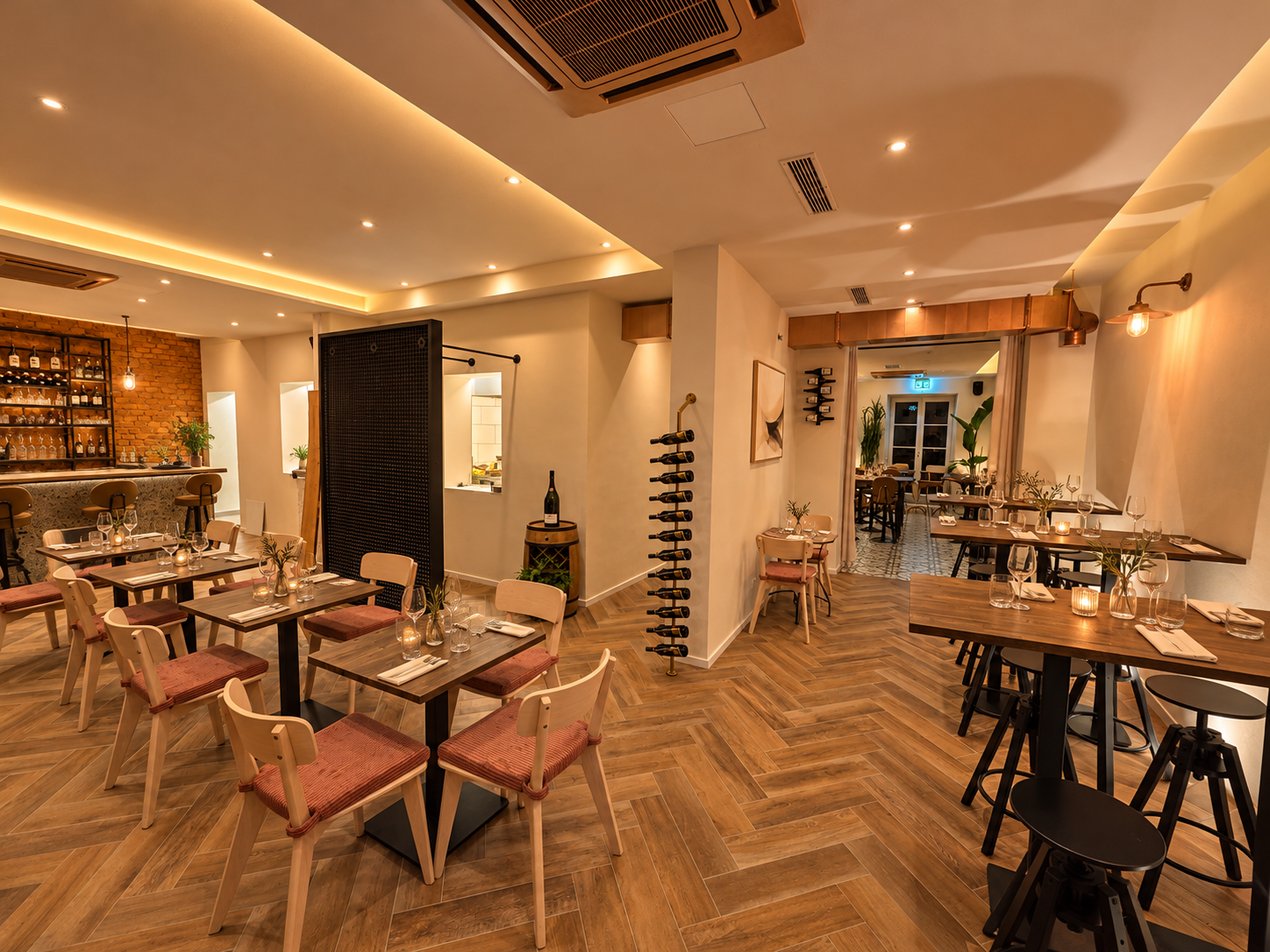 Warmly lit modern restaurant interior with wooden tables, upholstered chairs, bar, wine racks, and exposed brick walls