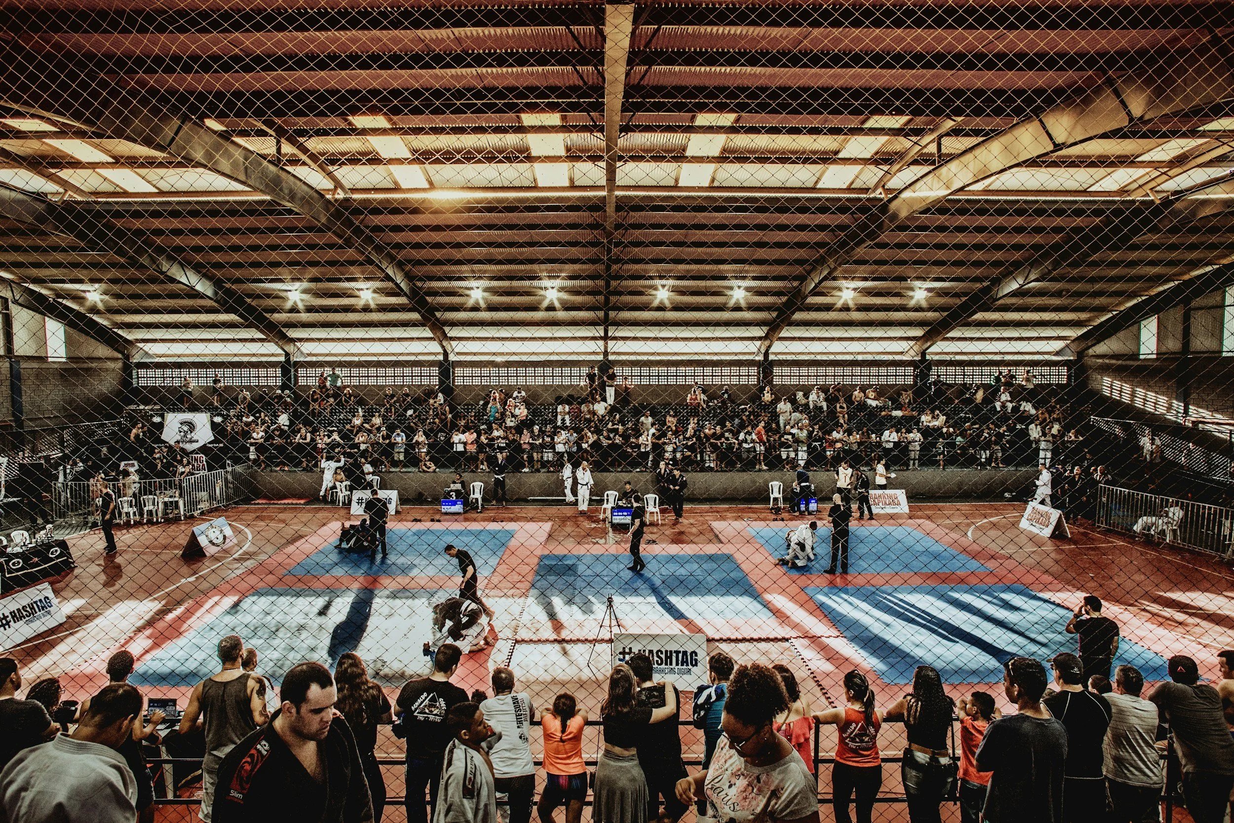 Crowded indoor Brazilian jiu-jitsu tournament with athletes competing on blue and red mats behind a chain-link fence