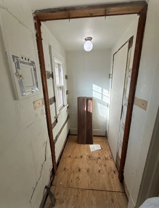 Narrow unfinished hallway under renovation with exposed doorframe, plywood floor, electrical panel, window, and cabinet.