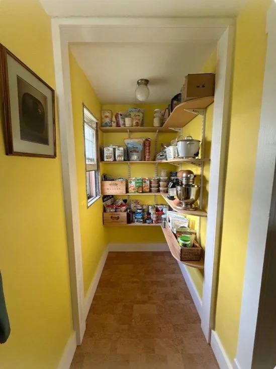 Narrow yellow walk-in pantry with open shelves stocked with canned goods, baking supplies, kitchen appliances, and storage boxes.