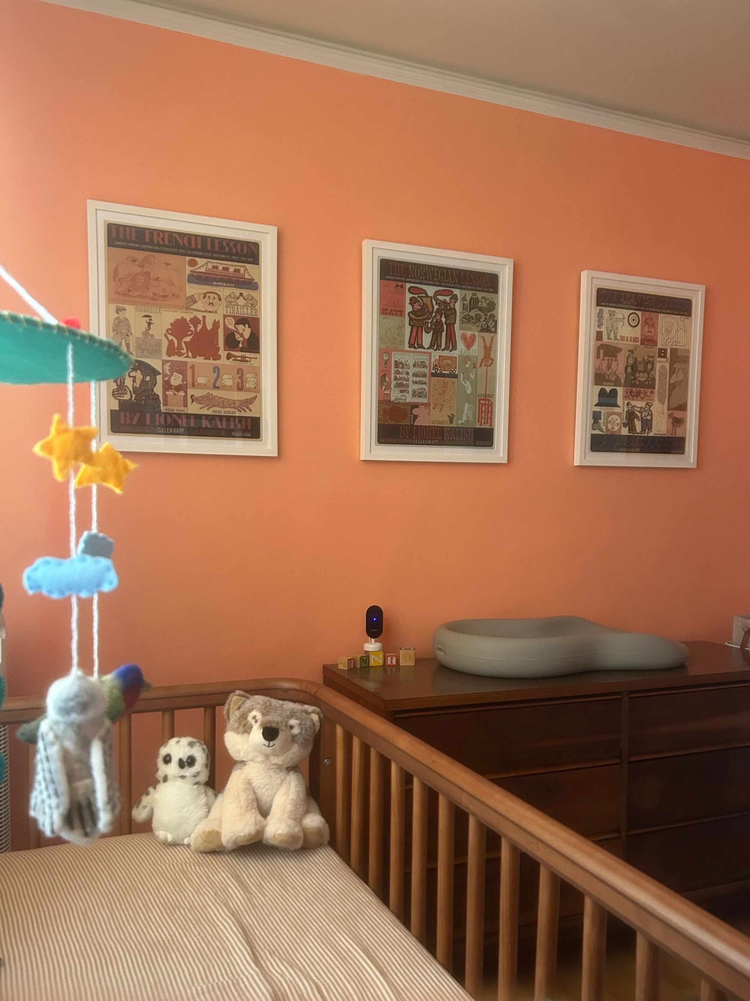 Nursery with three framed vintage posters on a coral wall, a wooden crib holding stuffed animals, and a dresser with a white baby monitor.