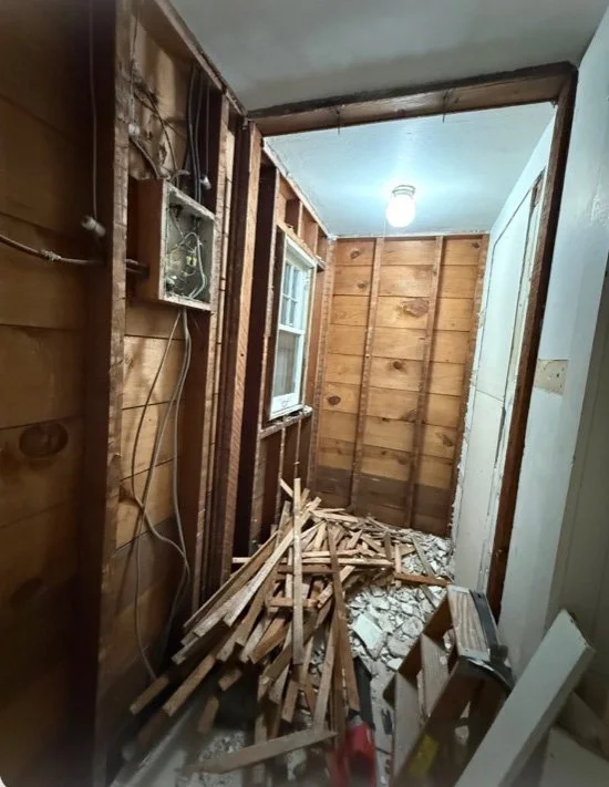 Narrow hallway under renovation with exposed wood framing, electrical wiring, debris, and a hanging light fixture