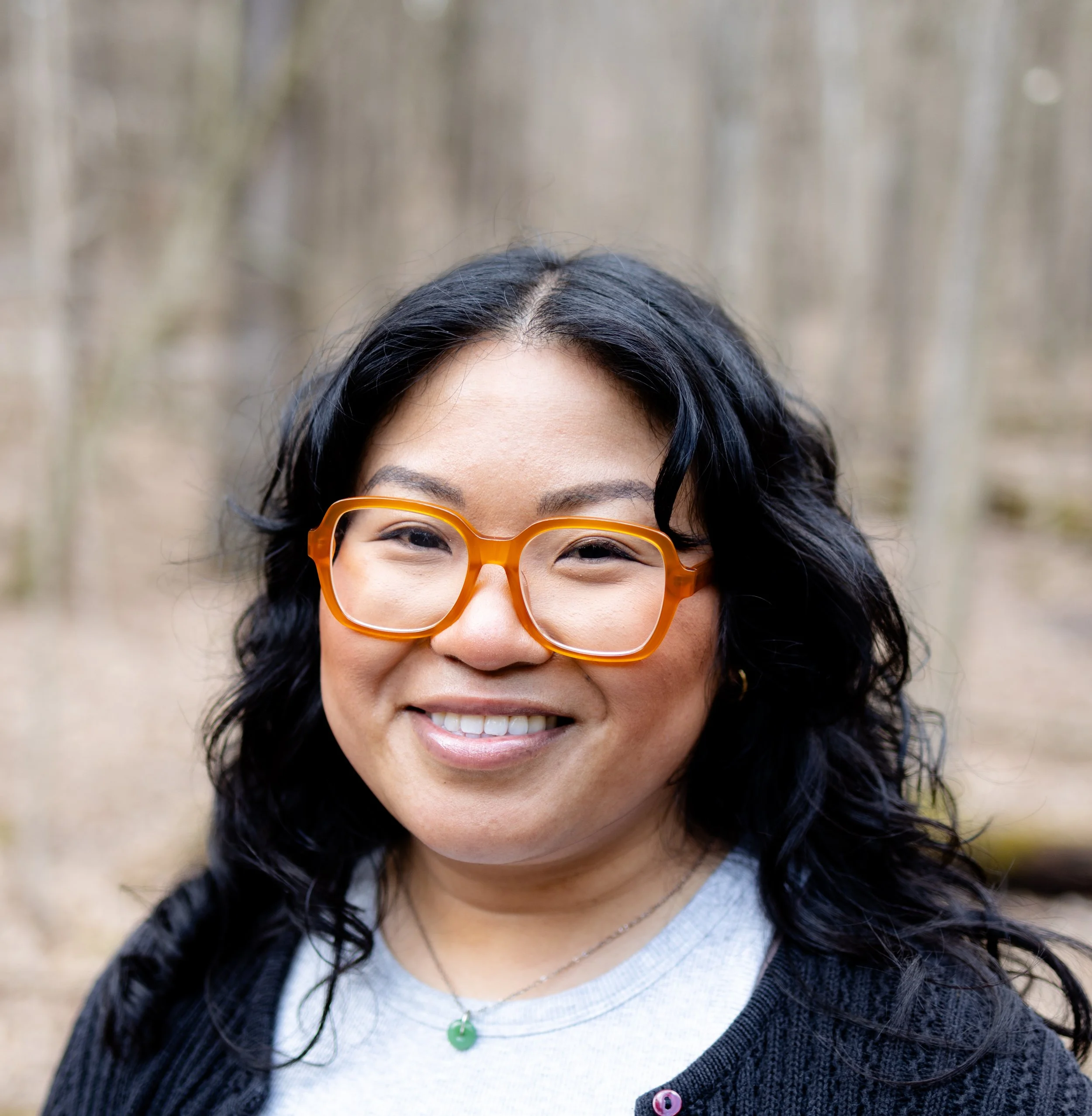 Smiling woman with black hair wearing oversized orange eyeglasses and a green pendant necklace